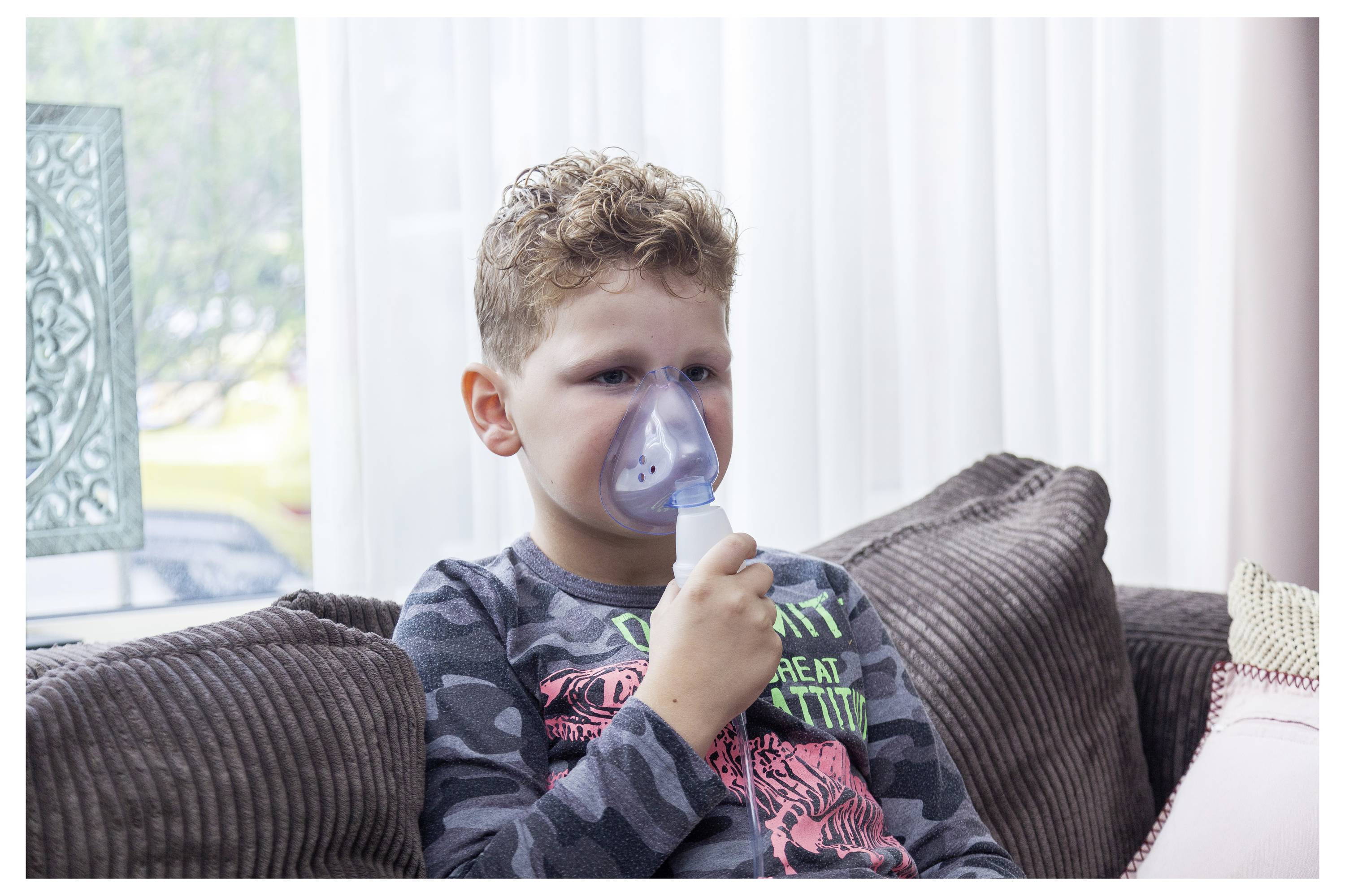 Un jeune garçon est assis sur un canapé avec un masque de nébuliseur pour un traitement respiratoire, ce qui suggère une préoccupation concernant la santé respiratoire ou une maladie.