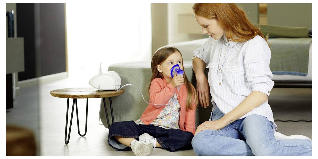 Une mère regarde son jeune enfant utiliser un nébuliseur à la maison, assis ensemble sur le sol. L'enfant inhale le médicament du dispositif.
