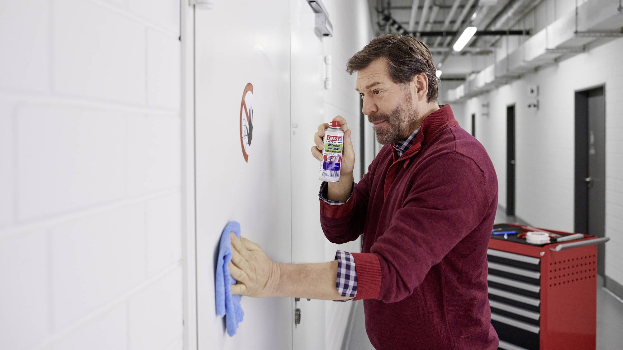 Un homme nettoie soigneusement un mur métallique avec un produit de nettoyage et un chiffon dans un couloir industriel. À l'arrière-plan, un chariot à outils est visible.