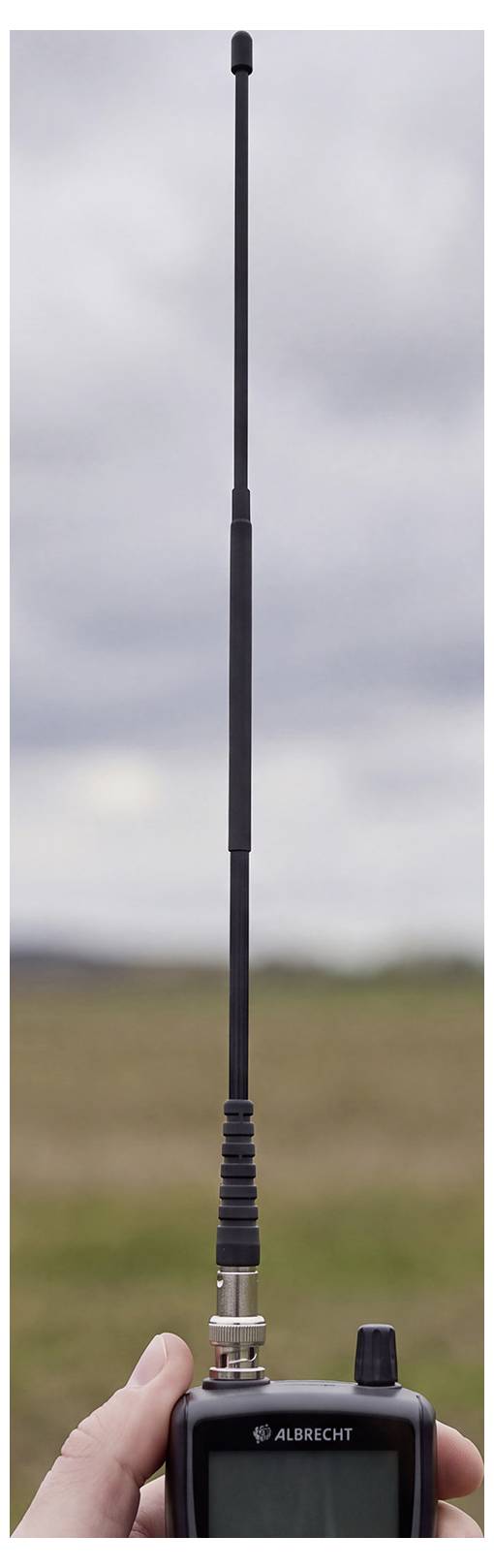 Radio bidirectionnel portable Albrecht avec une longue antenne verticale déployée contre un ciel nuageux extérieur et un arrière-plan de champ.