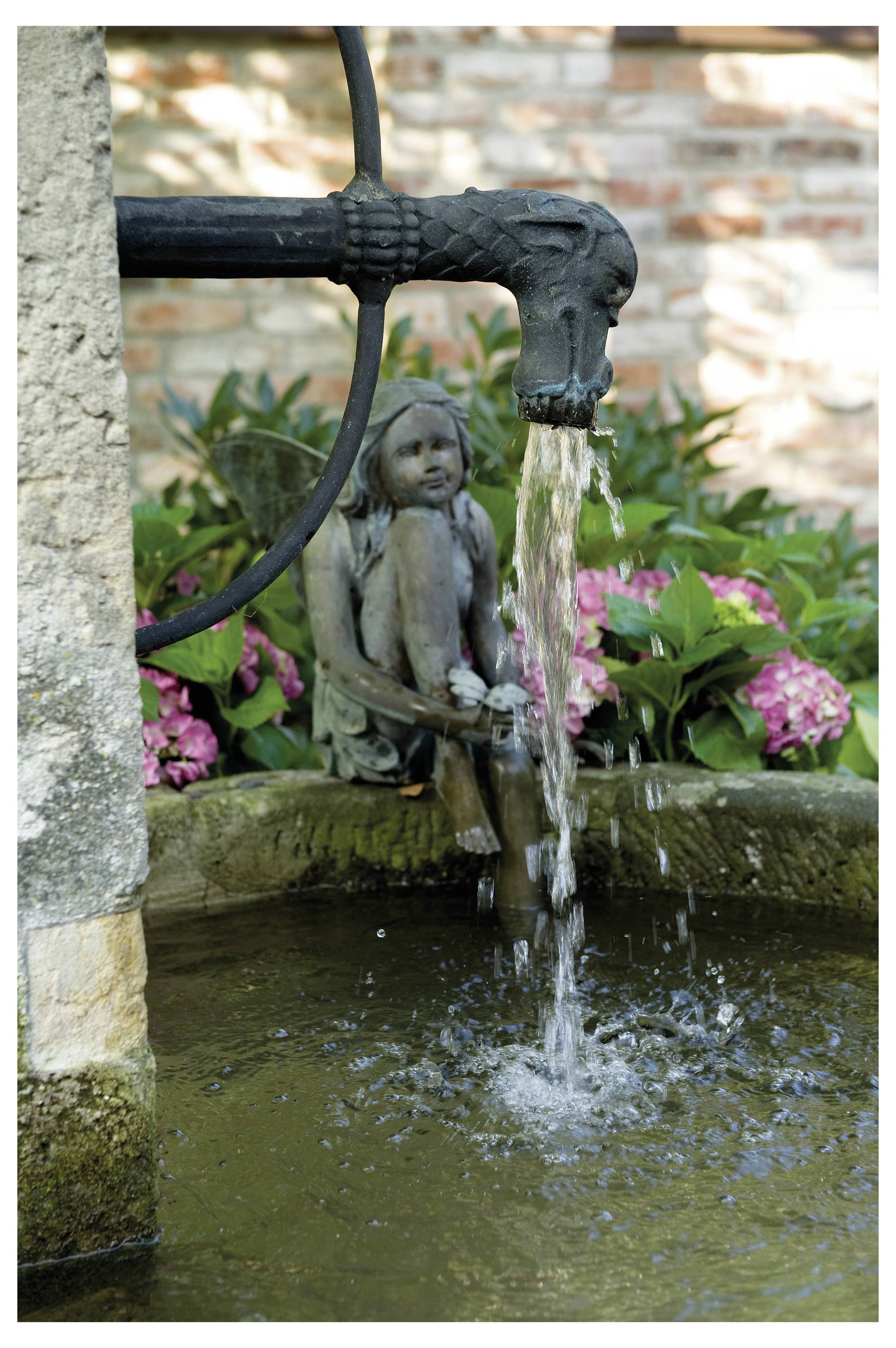 Une fontaine de pierre avec un déversoir en forme de dragon déverse de l'eau dans un bassin, entourée de verdure luxuriante et de fleurs roses, avec une statue de fée.