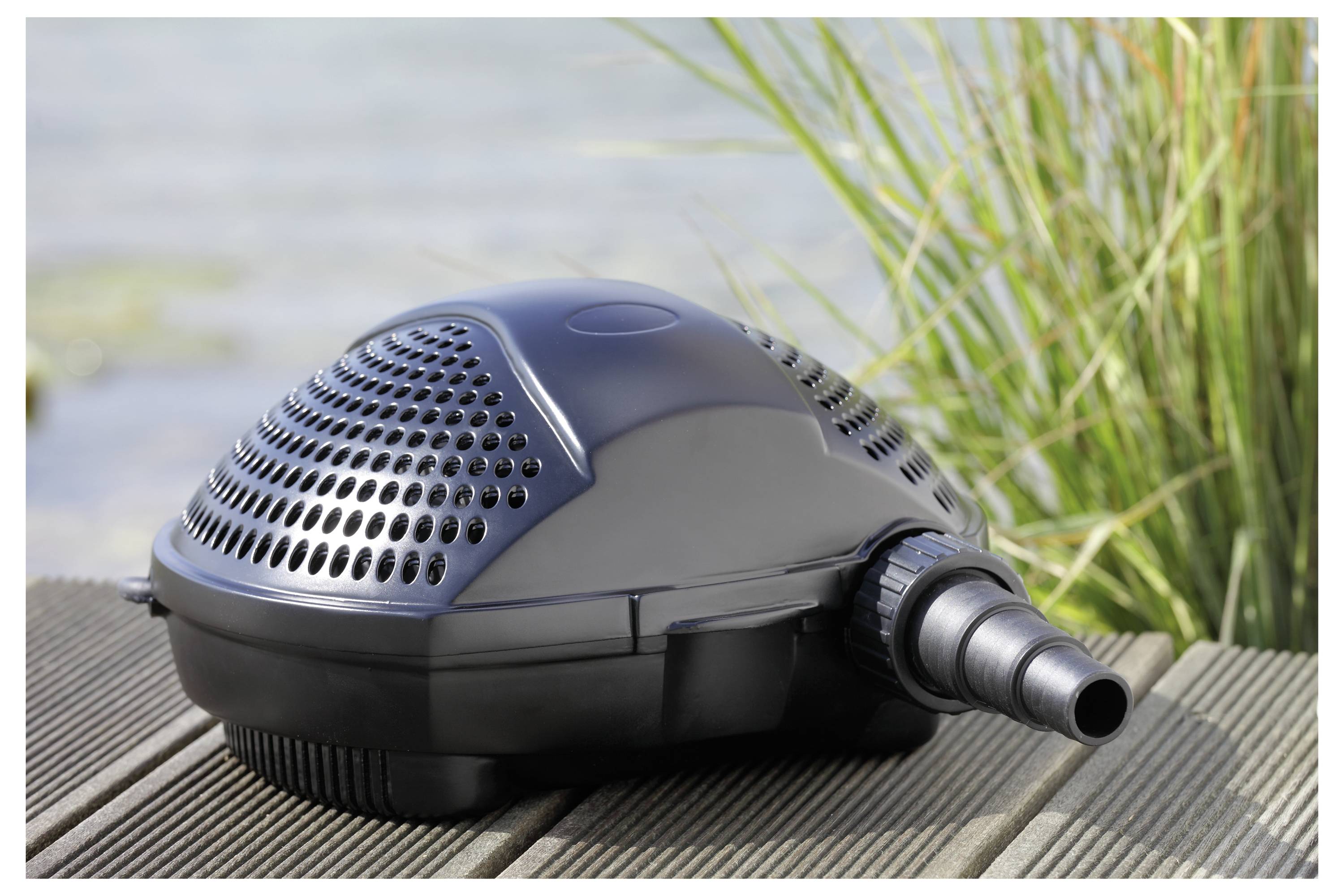 Une pompe de bassin noire placée sur un pont en bois près de l'eau et de hautes herbes, montrant sa vue de côté et son connecteur de tuyau.