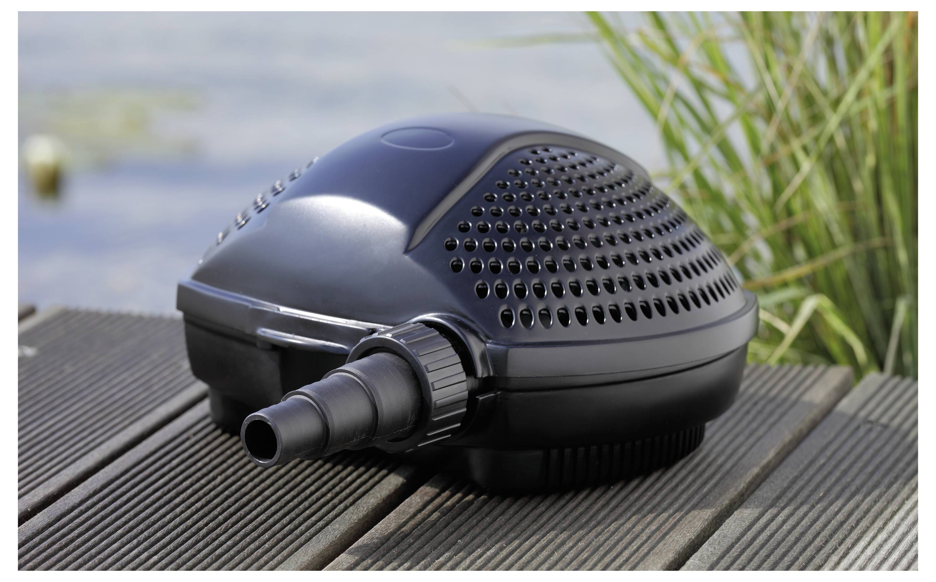 Une pompe noire pour bassin est posée sur une terrasse en bois près de l'eau et de l'herbe, présentant un design aéré et un raccordement de tuyau pour la circulation de l'eau.