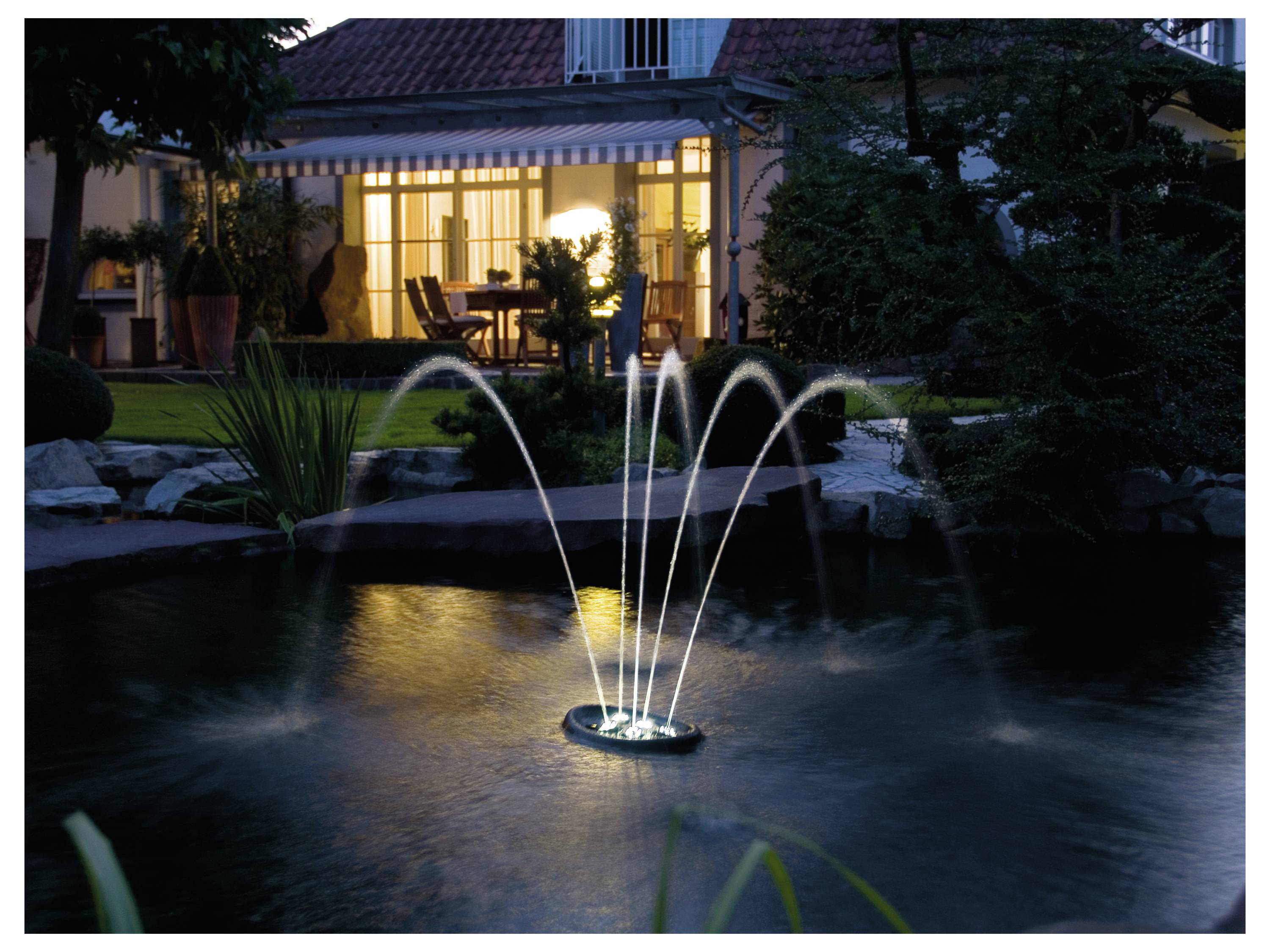 Une fontaine éclairée dans un bassin de jardin la nuit, avec l'intérieur chaleureusement illuminé d'une maison visible en arrière-plan, créant une atmosphère sereine.