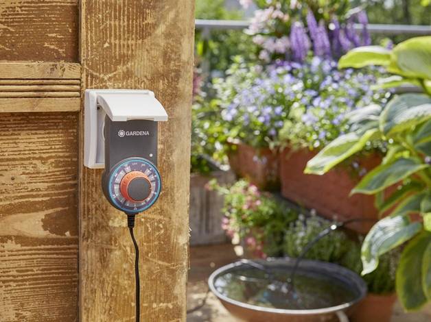 Un système d'irrigation automatique est installé sur une clôture en bois. En arrière-plan, des plantes en fleurs et une petite fontaine sont visibles.