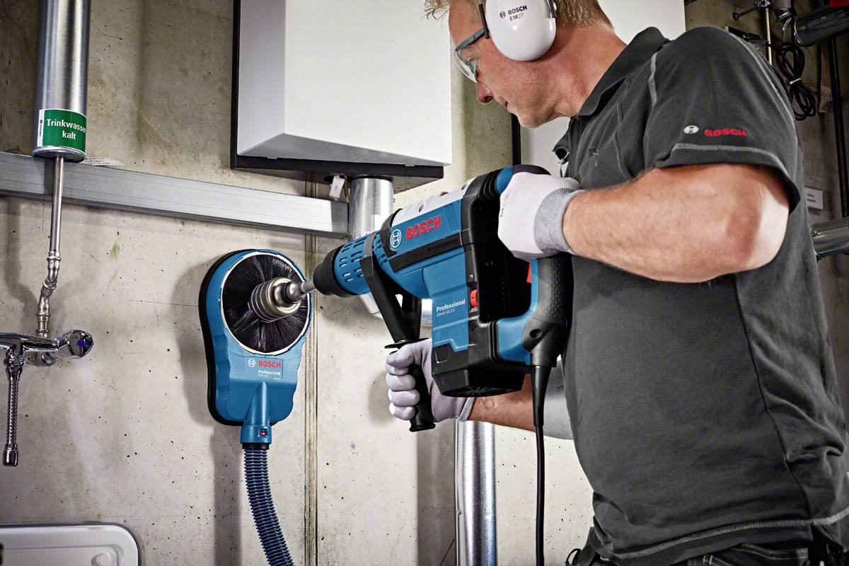 Un homme utilise une perceuse Bosch bleue avec un grand foret dans un bâtiment, portant des lunettes de protection et des protections auditives.