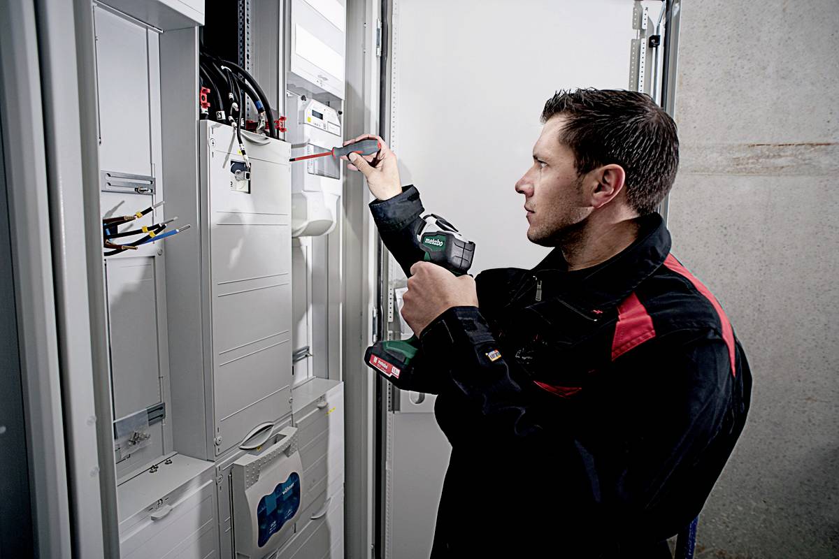 Un technicien vérifie un tableau électrique à l'aide d'un tournevis et d'un appareil de mesure. Il porte une veste de travail.