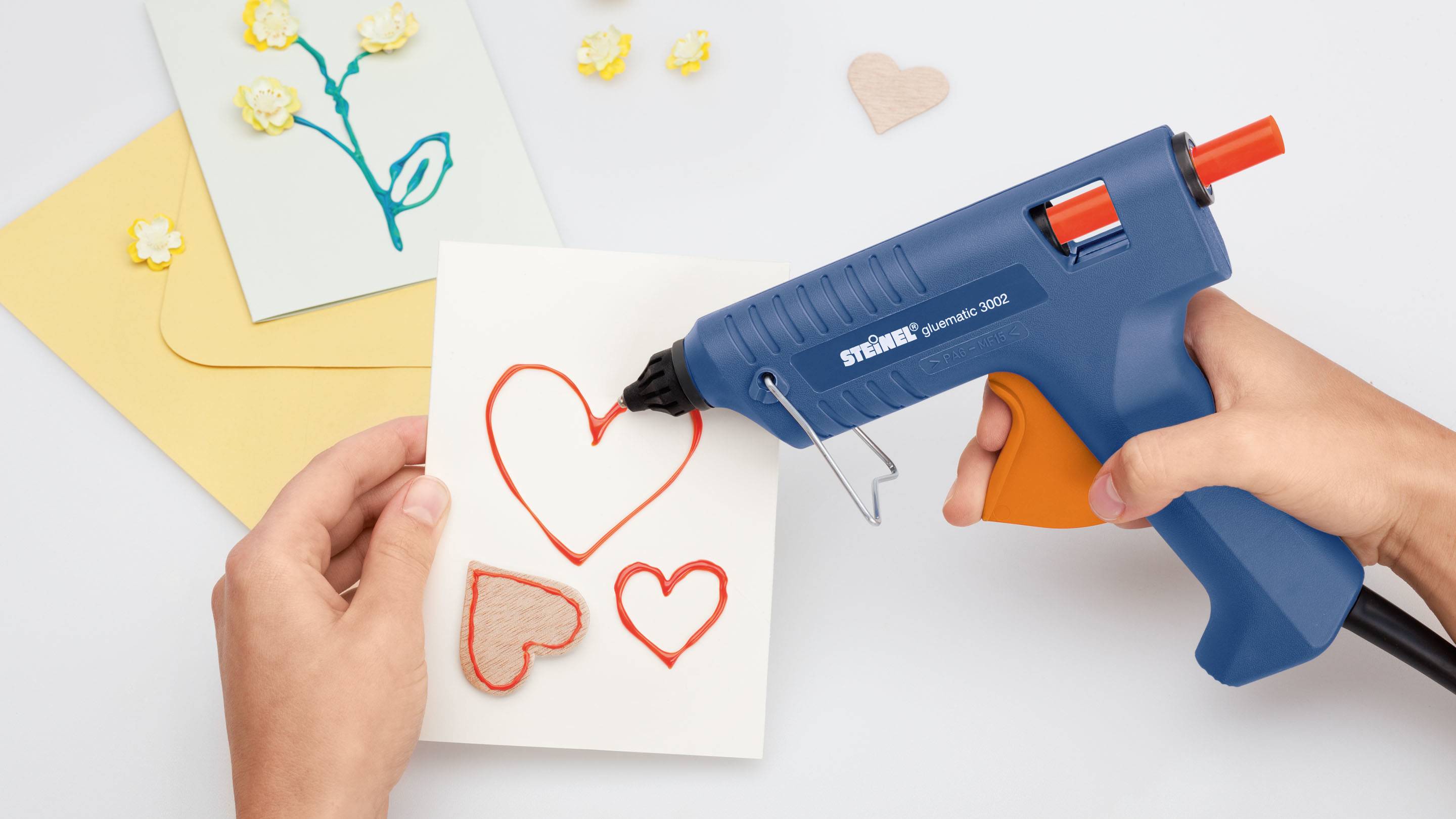 Une personne décore une carte avec des cœurs à l'aide d'un pistolet à colle chaude. D'autres cartes avec des motifs floraux sont visibles en arrière-plan.