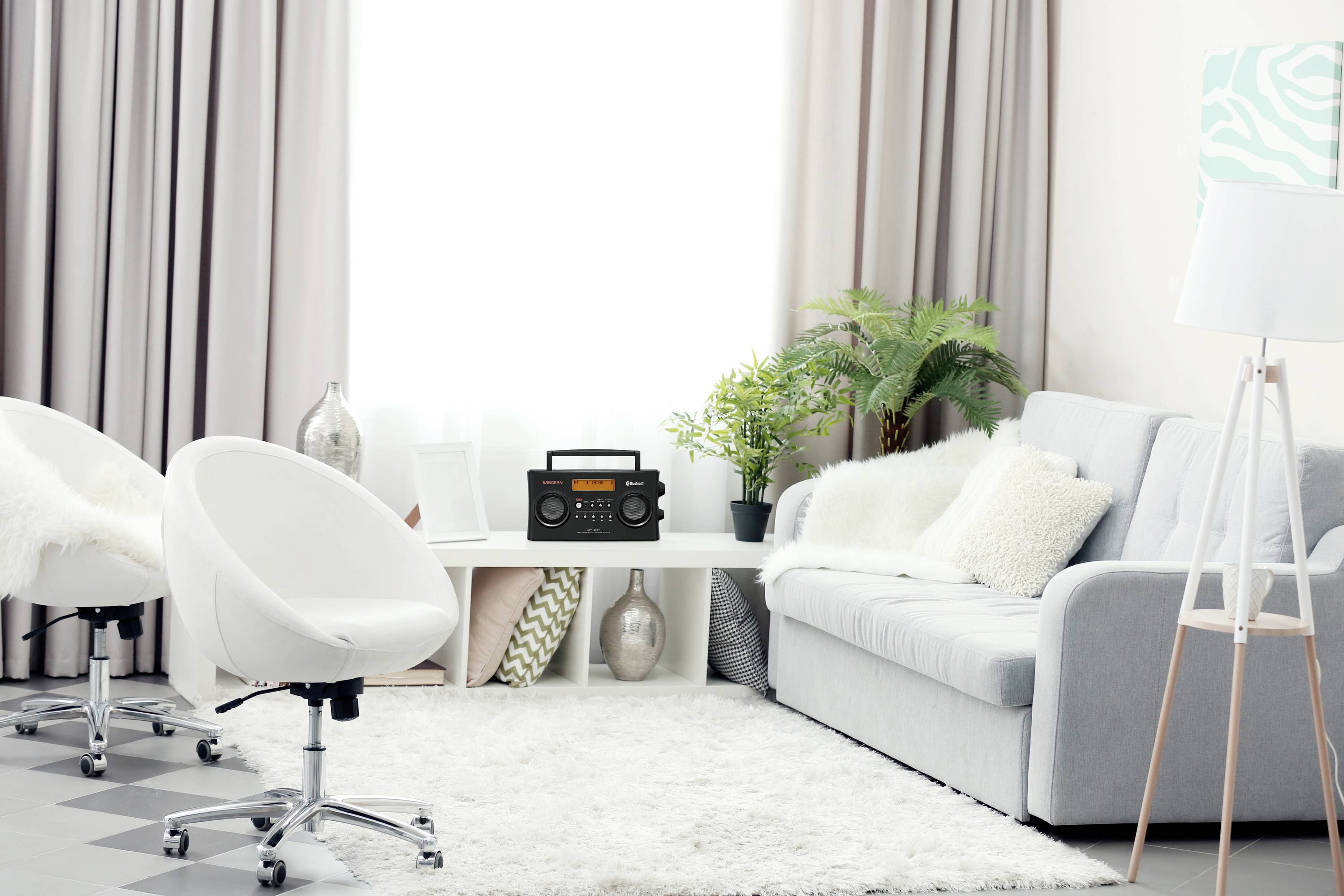 Salon lumineux avec un canapé gris, deux chaises blanches, une table basse avec une radio et des plantes devant une grande fenêtre dotée de rideaux.