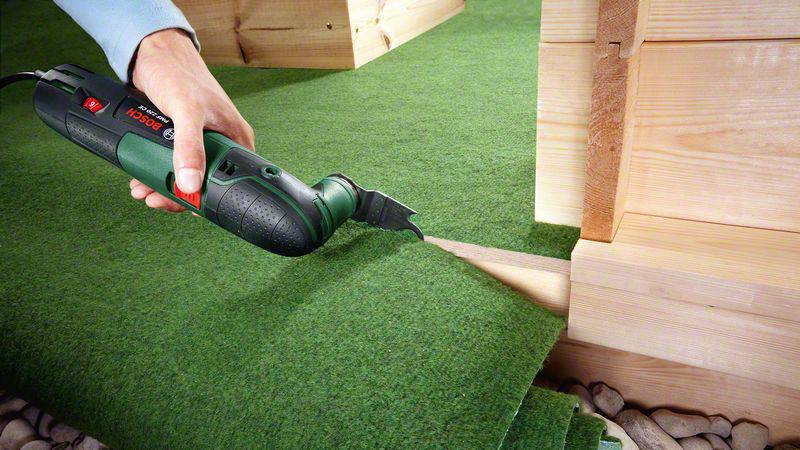 Une personne découpe un gazon artificiel avec un outil électrique pour l'ajuster autour du coin d'un bâtiment en bois.