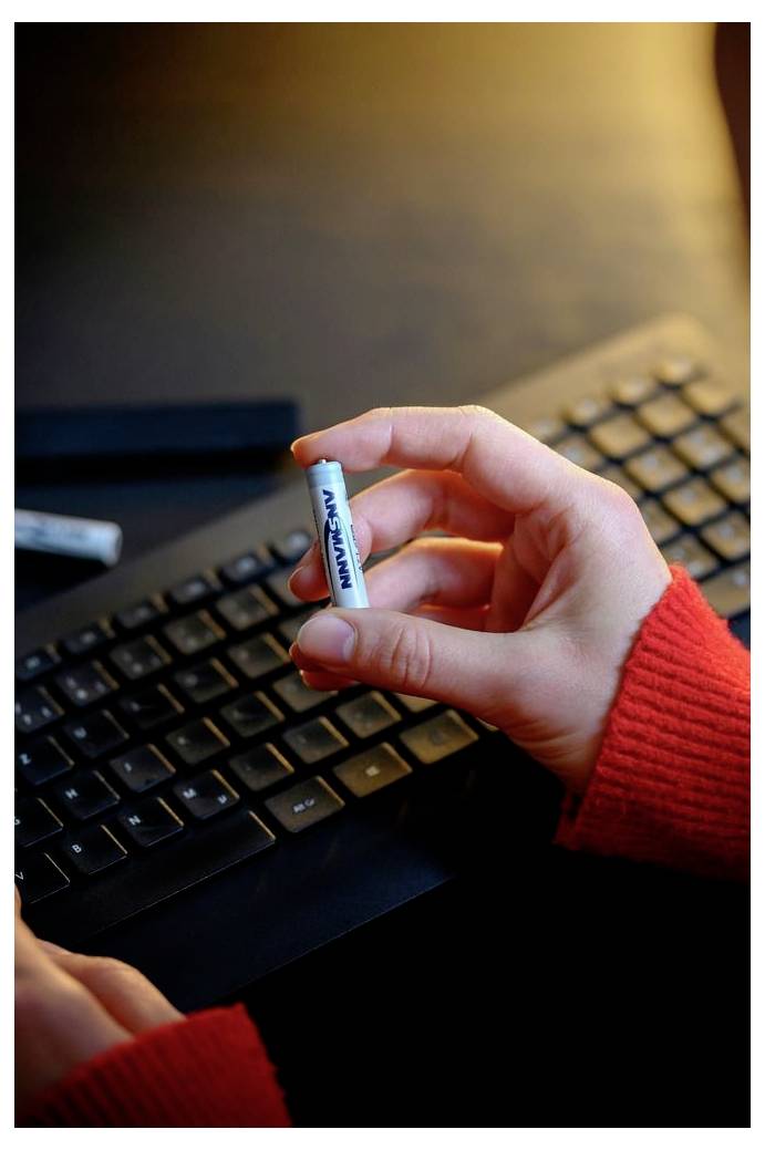 Une main tient une pile Panasonic AA au-dessus d'un clavier d'ordinateur noir, avec une manche de pull rouge visible.