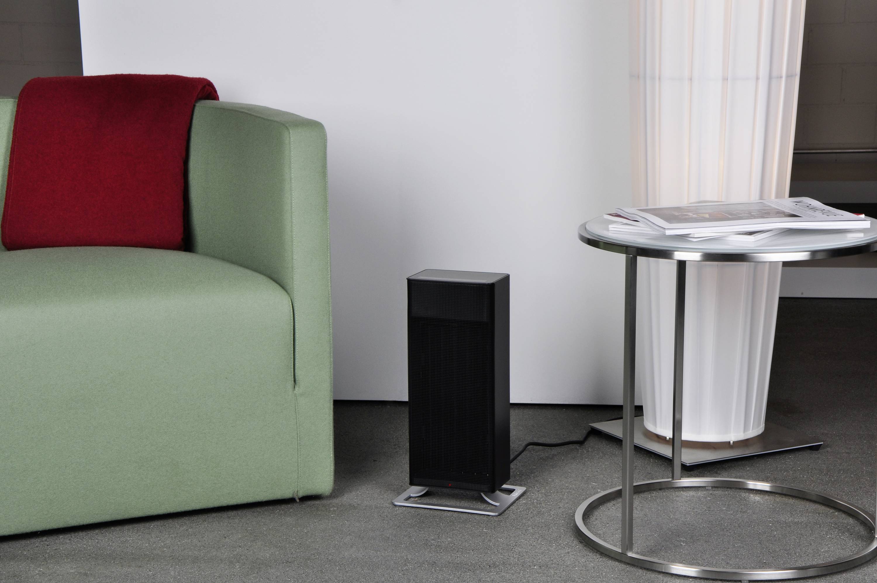 Un petit radiateur noir compact est placé à côté d'un fauteuil vert avec une couverture rouge et une table basse ronde sur laquelle sont posés des journaux.