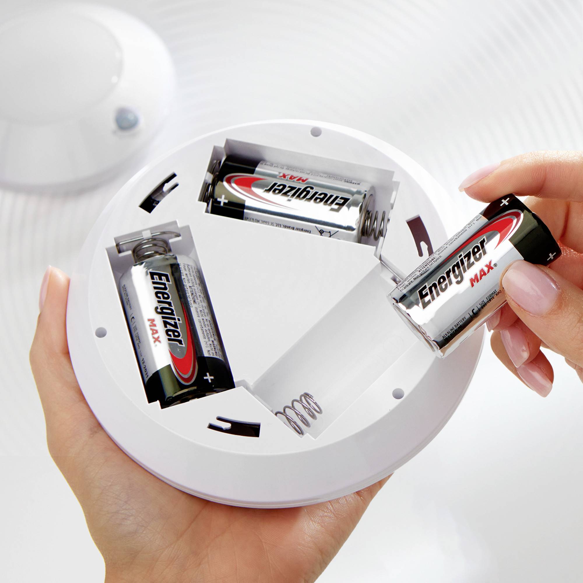 Deux mains tiennent un détecteur de fumée dans lequel des piles sont en cours d'insertion. Les piles, partiellement retirées, portent le logo 'Energizer'.