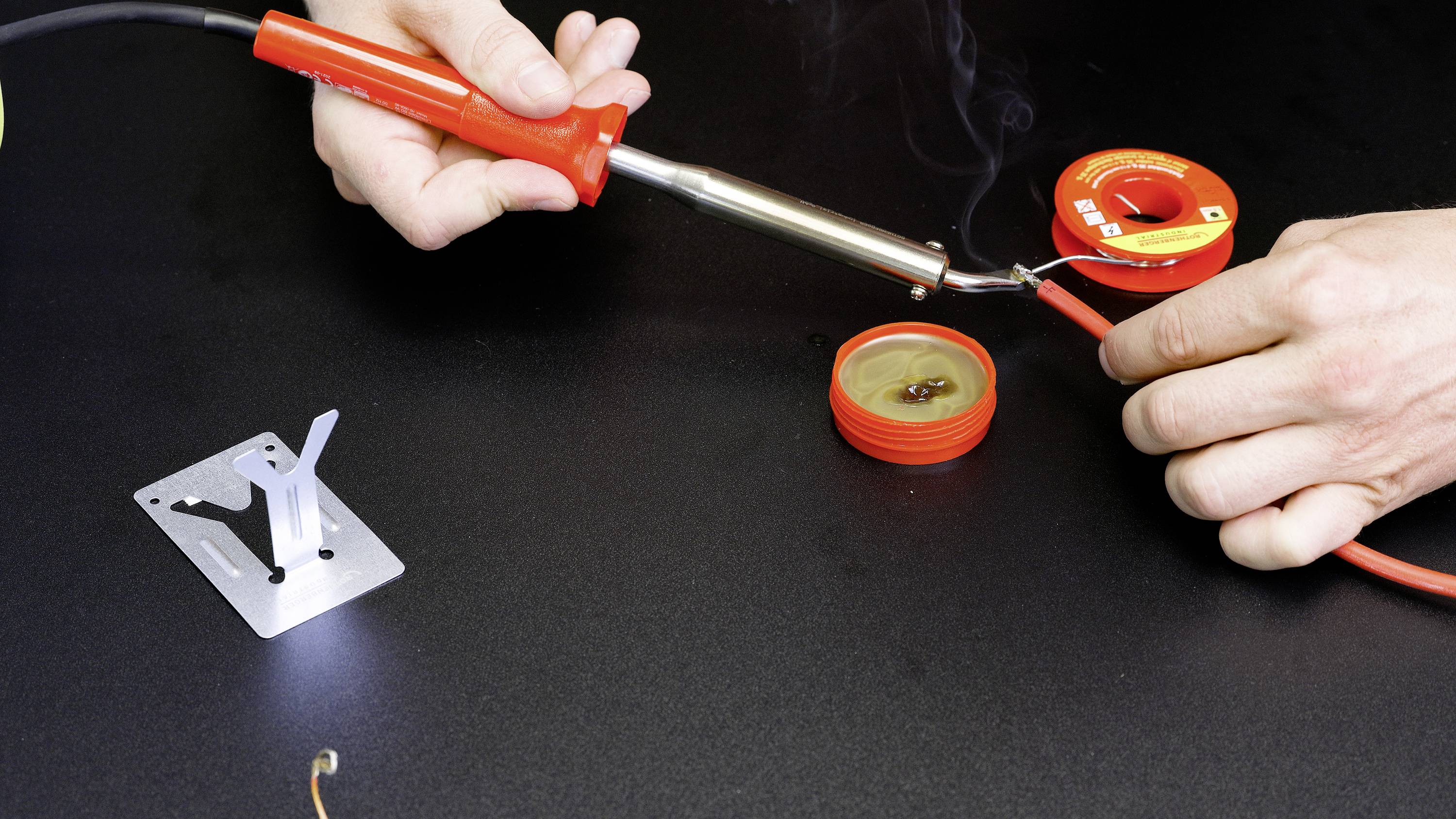 Une personne soude un câble avec un fer à souder. À côté du câble se trouvent de la soudure et des accessoires de soudage sur une table noire.