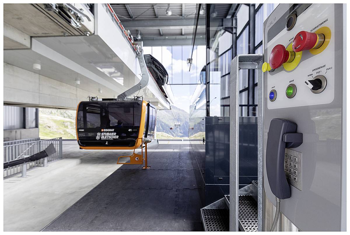 Station de téléphérique intérieure avec une télécabine orange, console de commande avec boutons au premier plan. Paysage montagneux visible à travers la fenêtre.