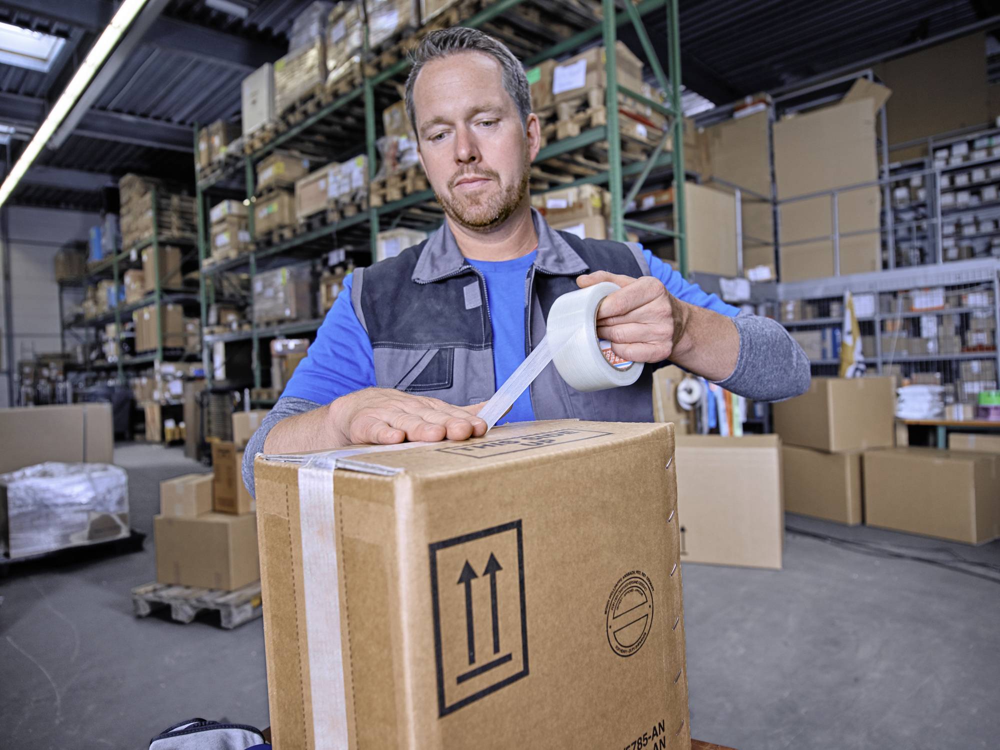 Un homme emballe un carton avec du ruban adhésif dans un entrepôt. Des étagères avec d'autres colis sont visibles en arrière-plan.