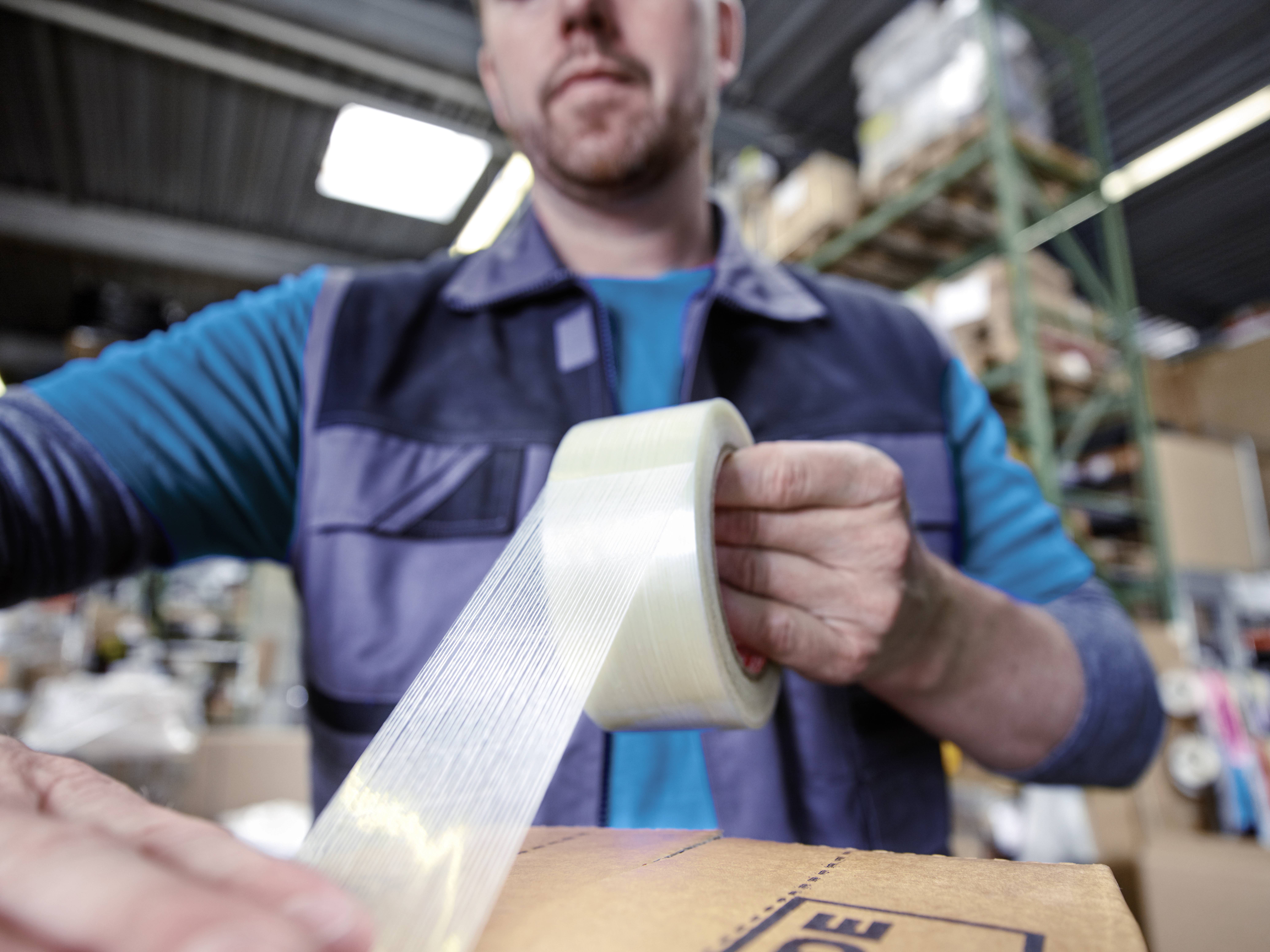 Un homme emballe un carton avec du ruban adhésif dans un environnement d'entrepôt. Des étagères avec des marchandises sont visibles en arrière-plan.