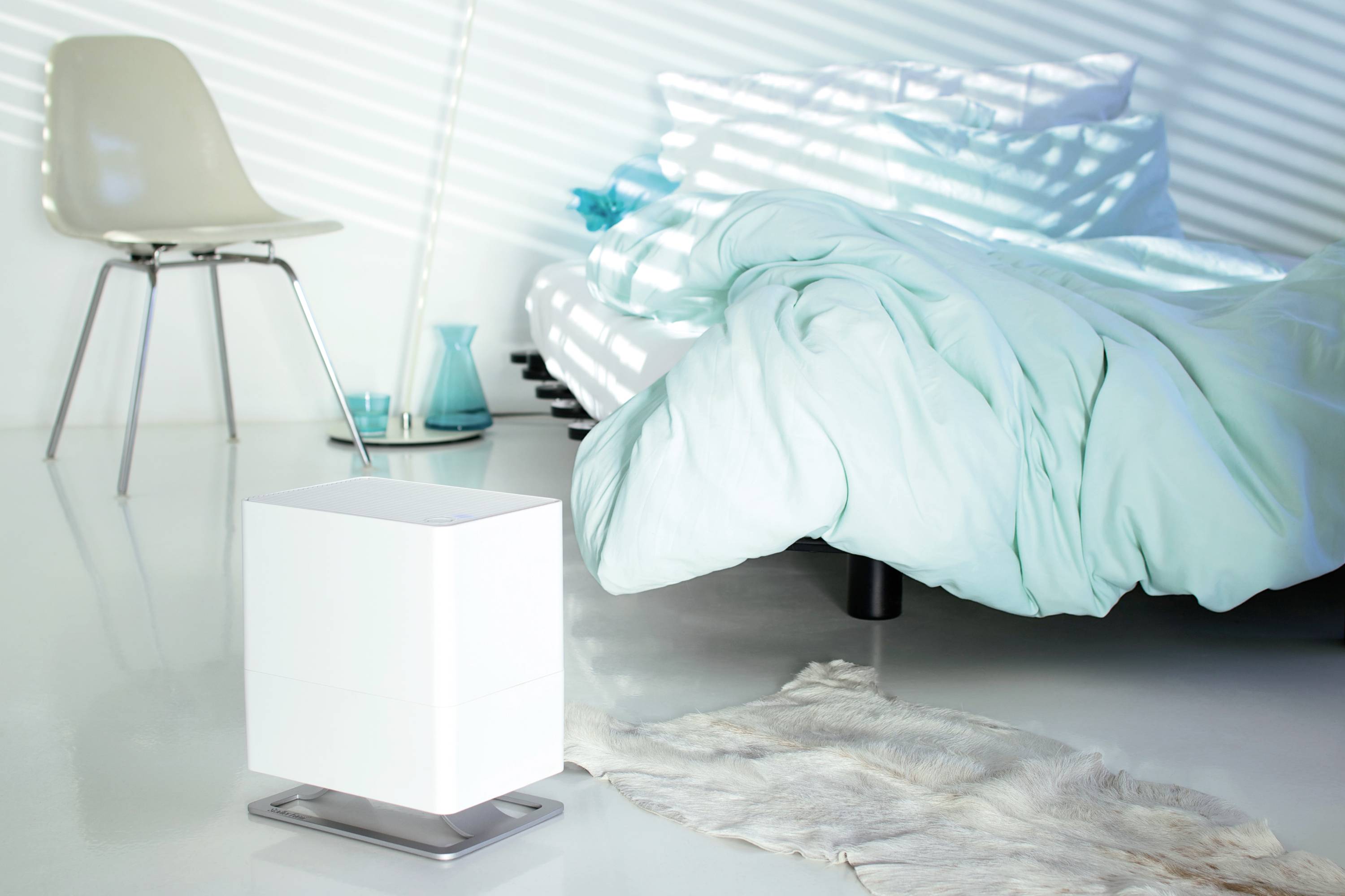 Un espace lumineux et moderne avec un humidificateur blanc posé sur le sol, une chaise blanche à gauche et un lit avec une couverture turquoise à droite.