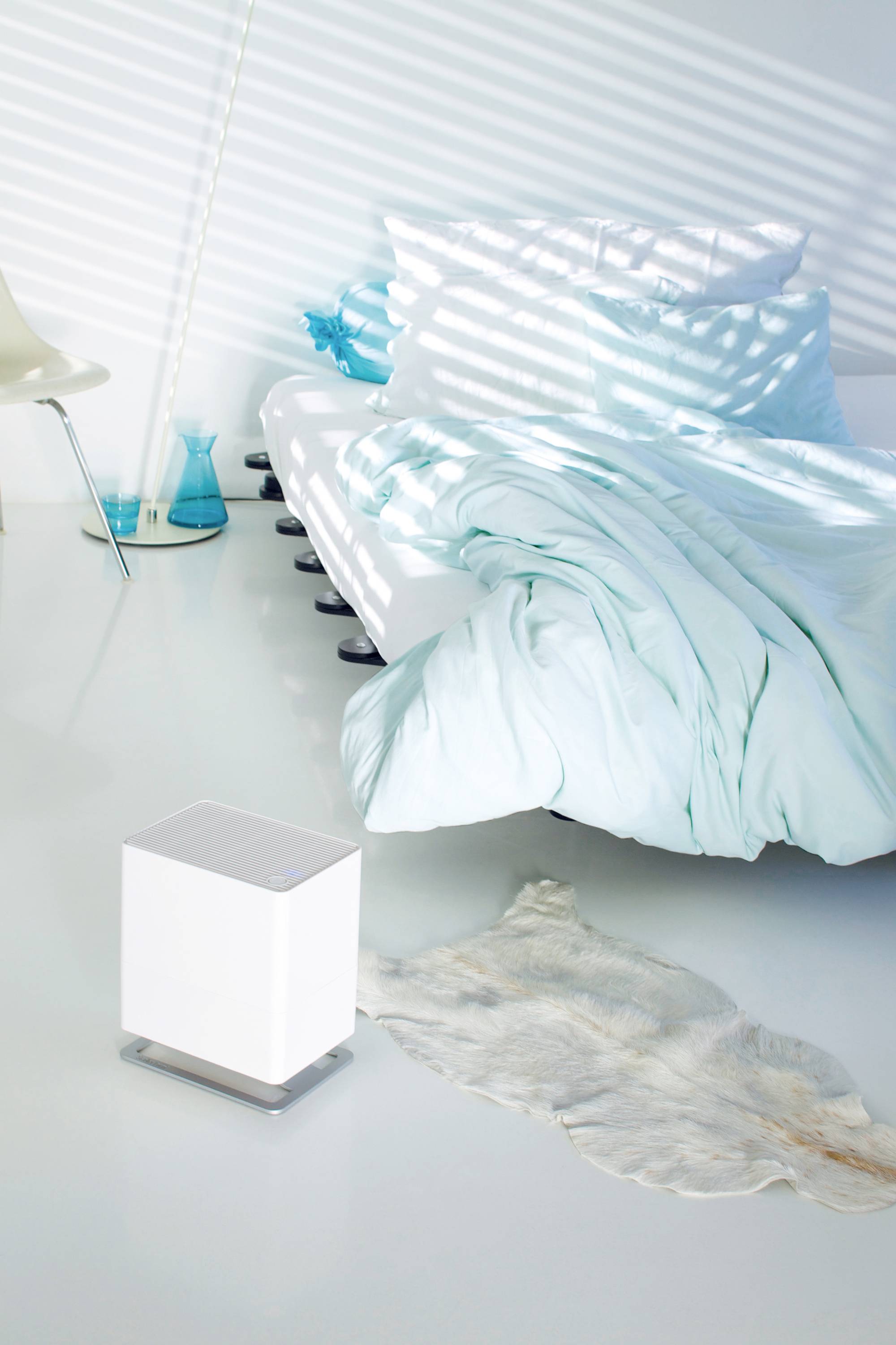 Une chambre à coucher avec un lit recouvert de draps de lit turquoise. À côté du lit, un purificateur d'air blanc est placé sur un tapis doux.