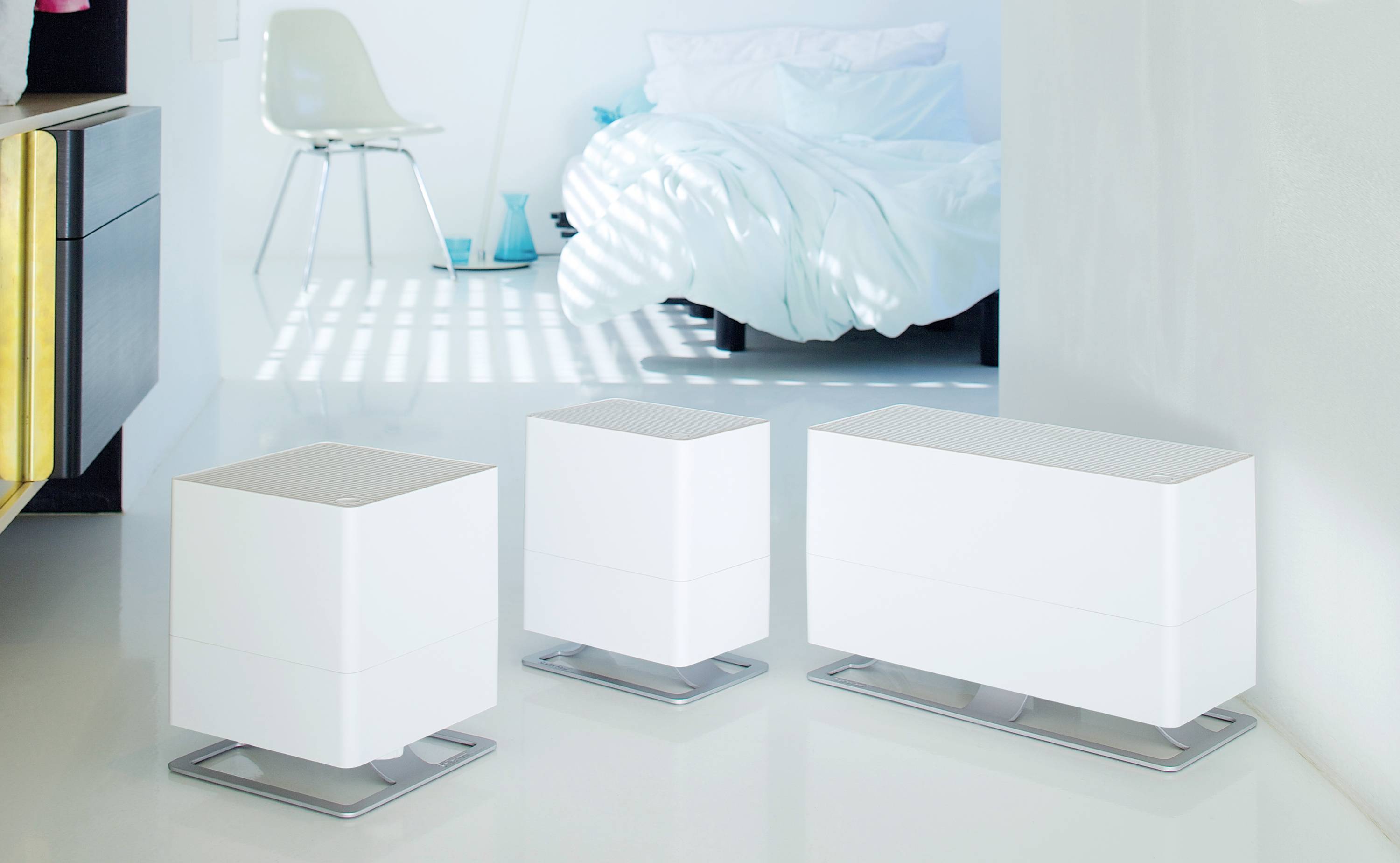Trois humidificateurs blancs modernes de différentes tailles sont placés dans une pièce au style minimaliste épuré, avec un lit en arrière-plan.