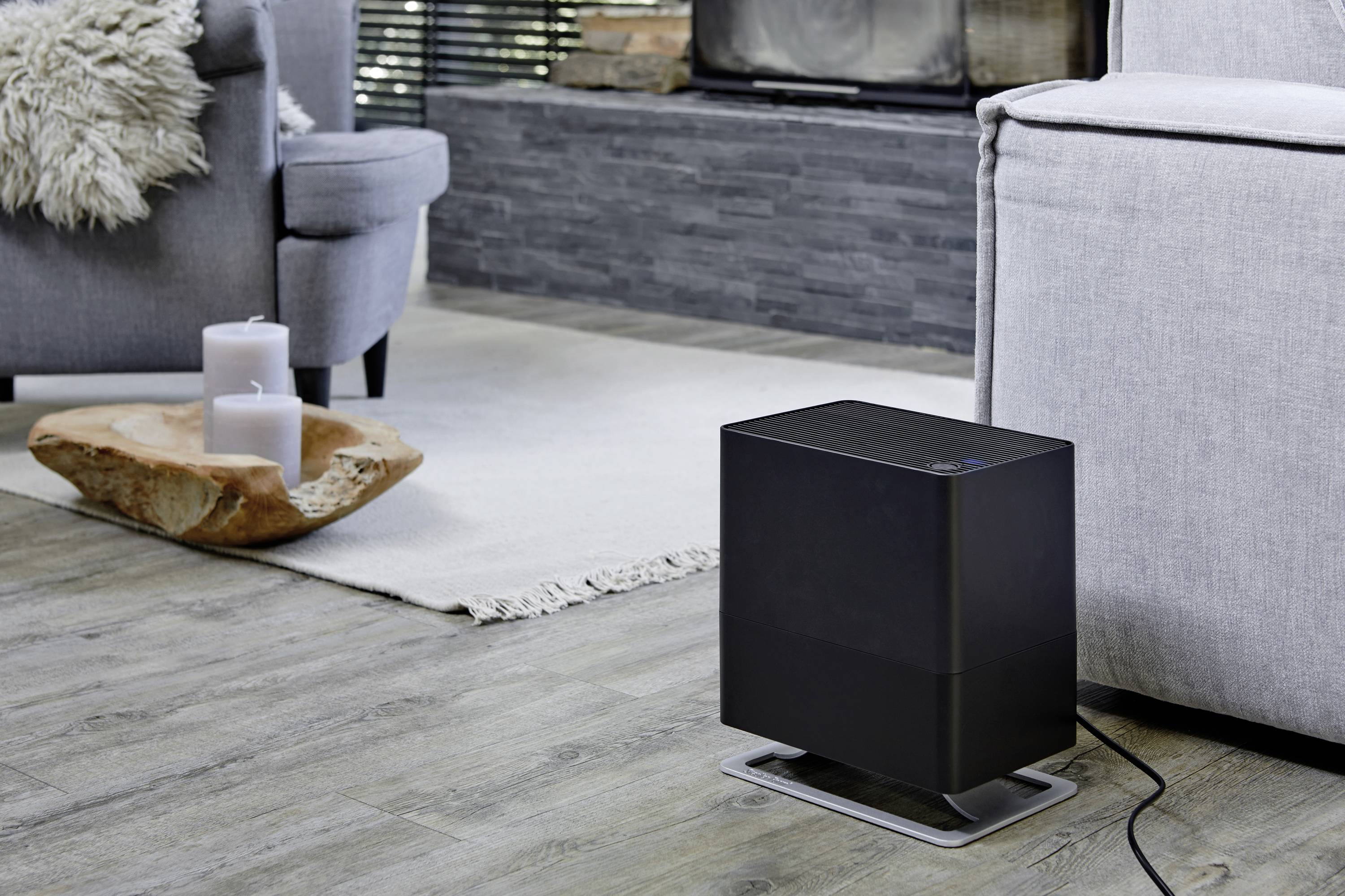 Un chauffage électrique noir est posé sur un sol en bois à côté d'un canapé gris dans un salon moderne. Des bougies sont placées dans un plateau en bois à proximité.