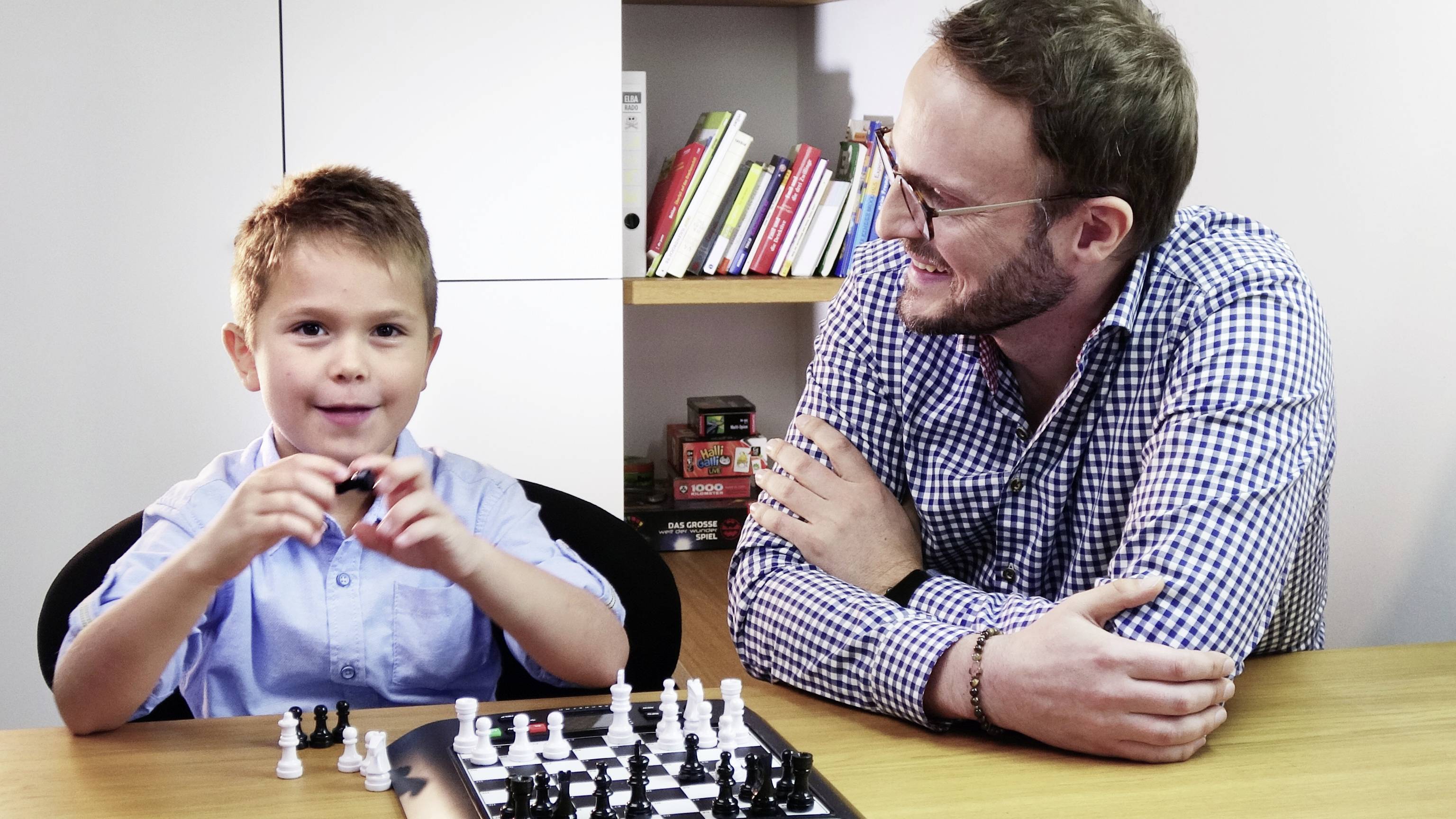 Un homme et un garçon sont assis à une table et jouent aux échecs. Le garçon tient une pièce dans sa main et sourit, tandis que l'homme le regarde avec bienveillance.