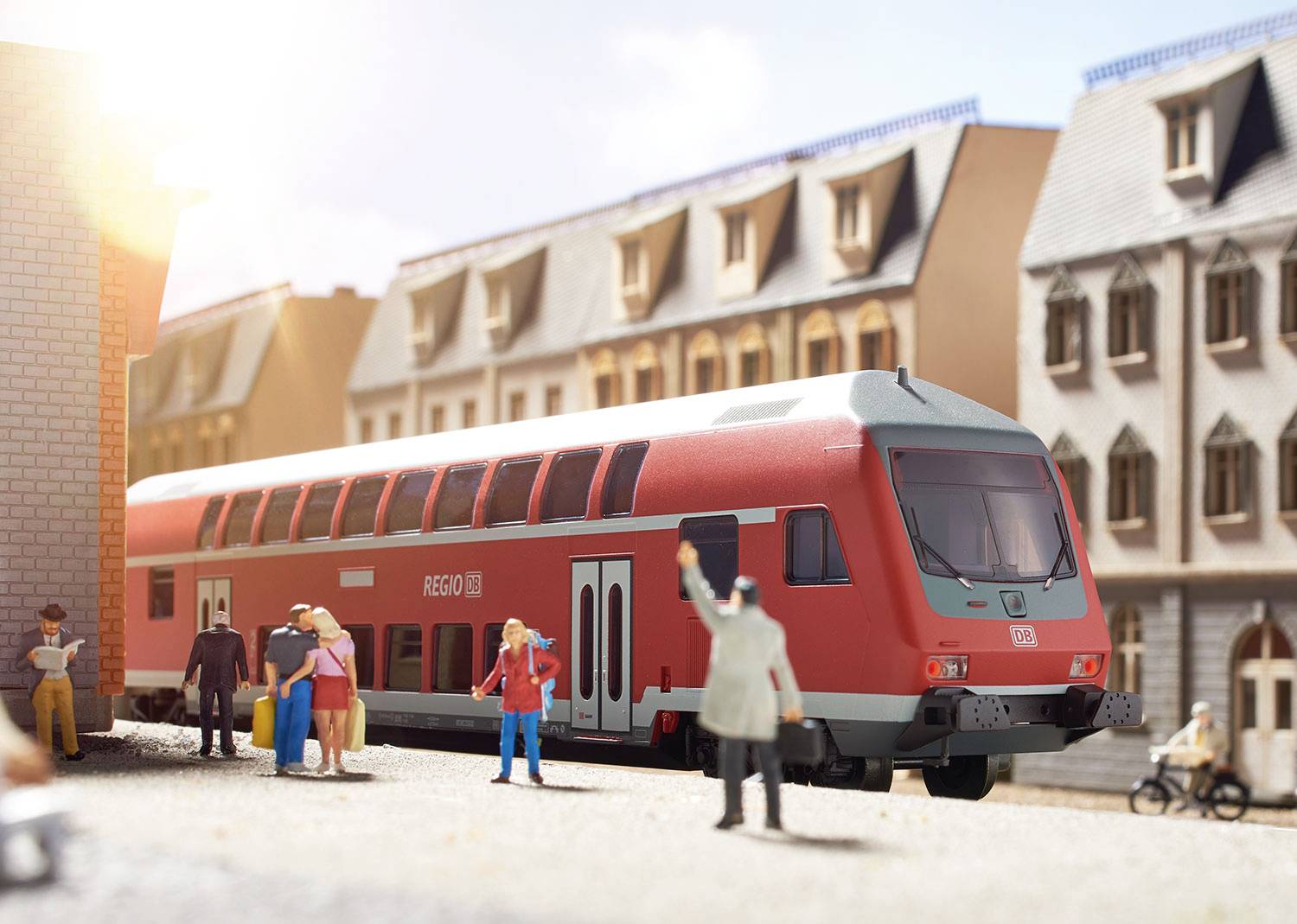 Un train régional rouge est stationné en gare, à côté de vieux bâtiments. Des personnes avec des bagages se déplacent sur le quai. Il fait soleil.