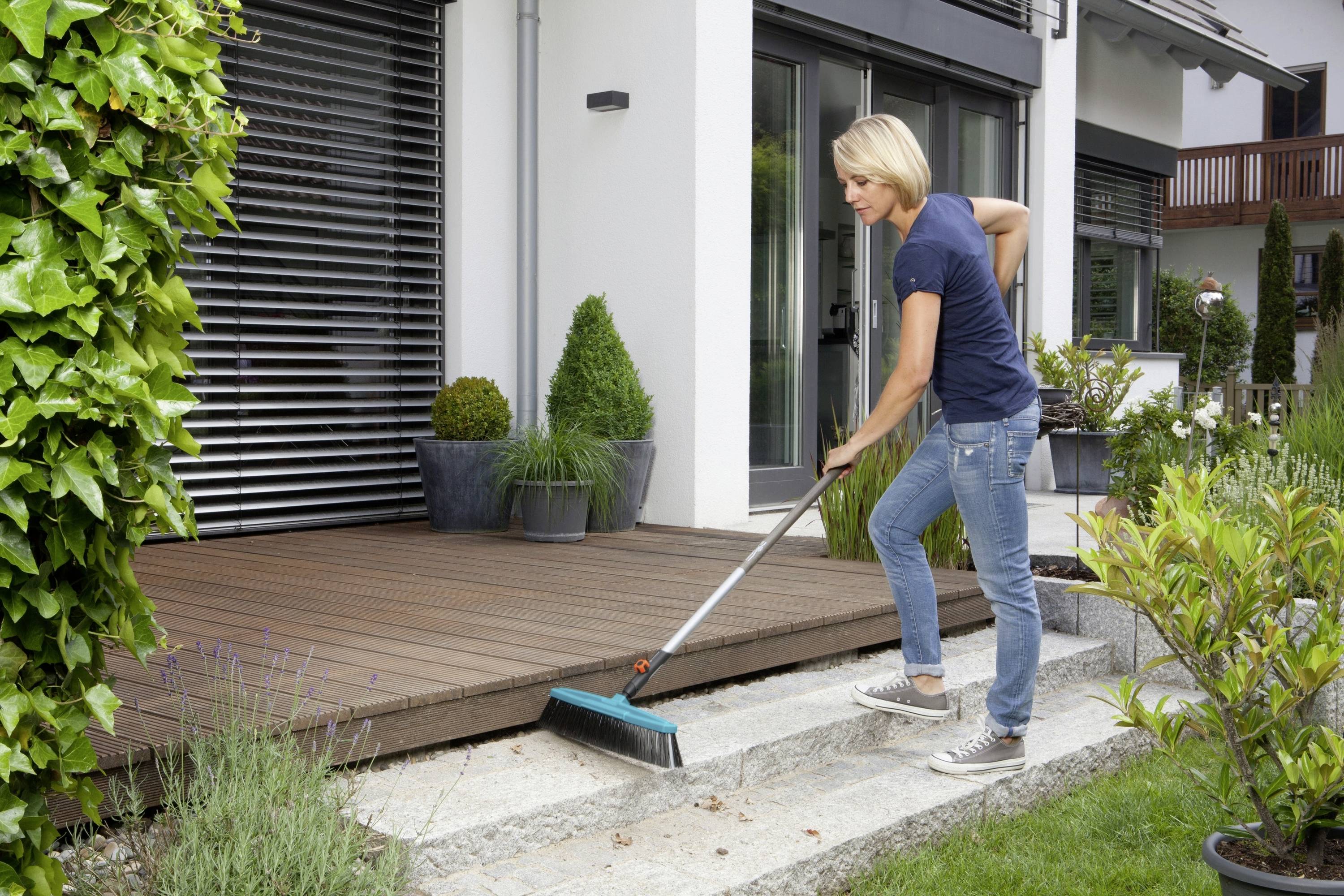 Une personne balaie l'escalier en pierre devant une terrasse. Des plantes et une maison moderne sont visibles en arrière-plan.