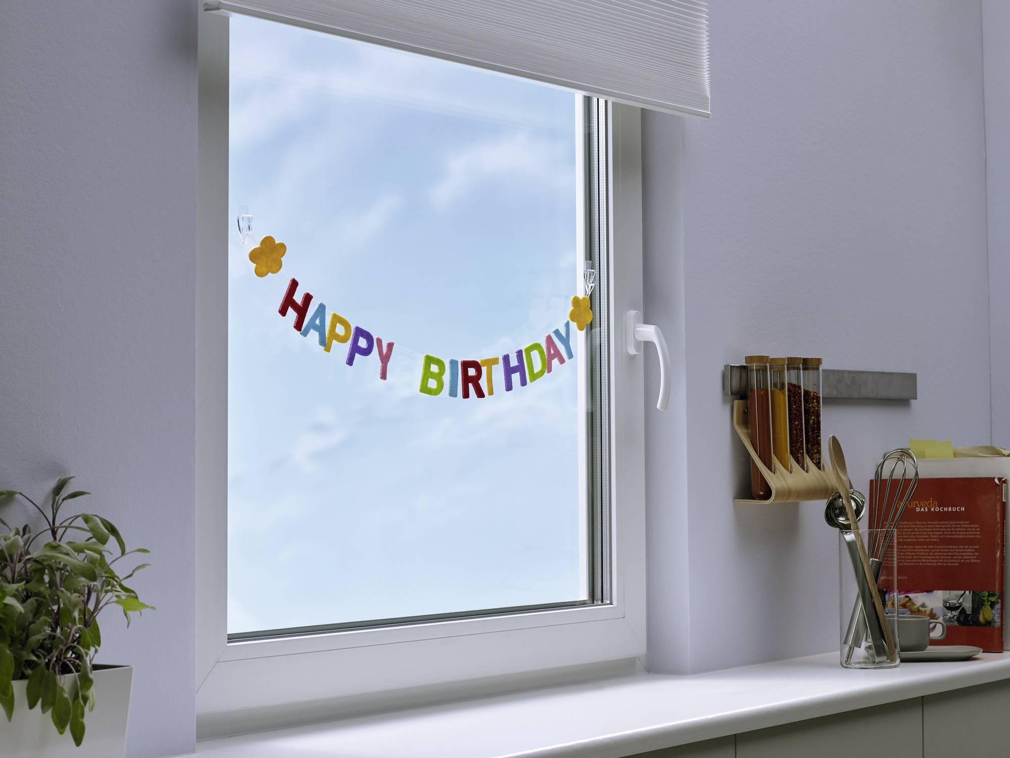 Décoration de fenêtre avec un message coloré 'Joyeux Anniversaire'. Ciel bleu et nuages en arrière-plan. Une plante en pot à gauche.