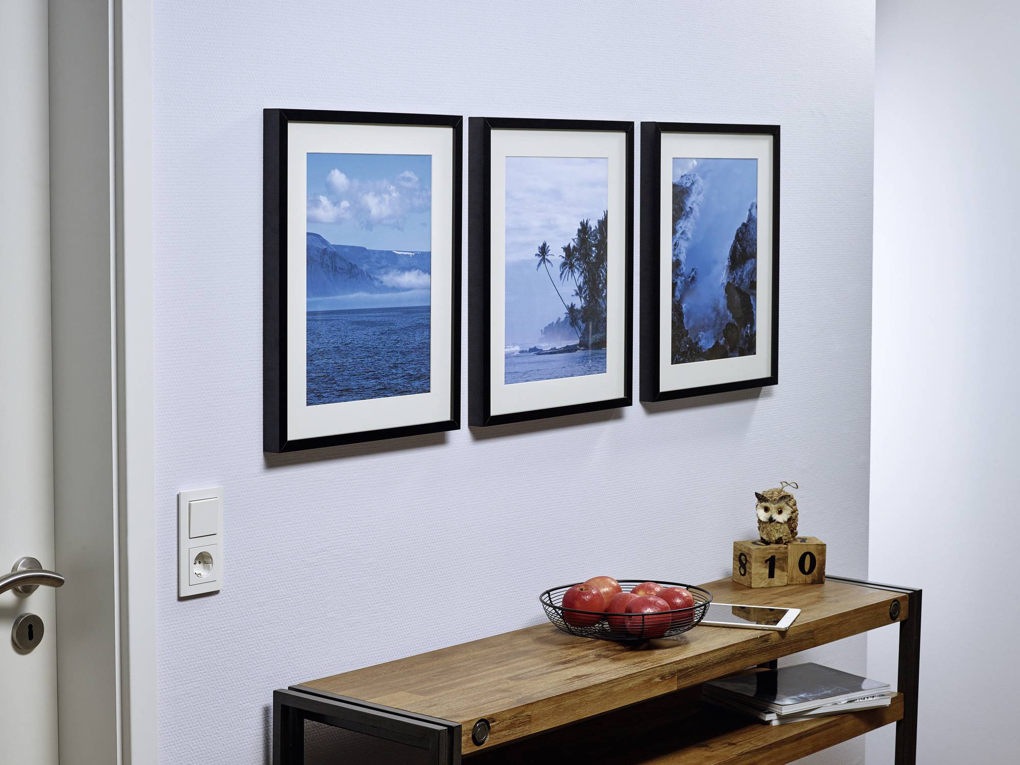 Trois photographies encadrées de paysages accrochées au mur au-dessus d'une table en bois portant un panier de fruits et une petite horloge surmontée d'une figurine de hibou.