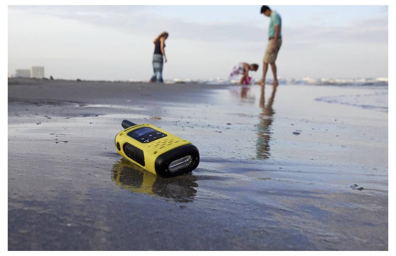 Talkie-walkie jaune portatif posé dans le sable mouillé de la plage, avec des personnes floues en arrière-plan près de la ligne de rivage.