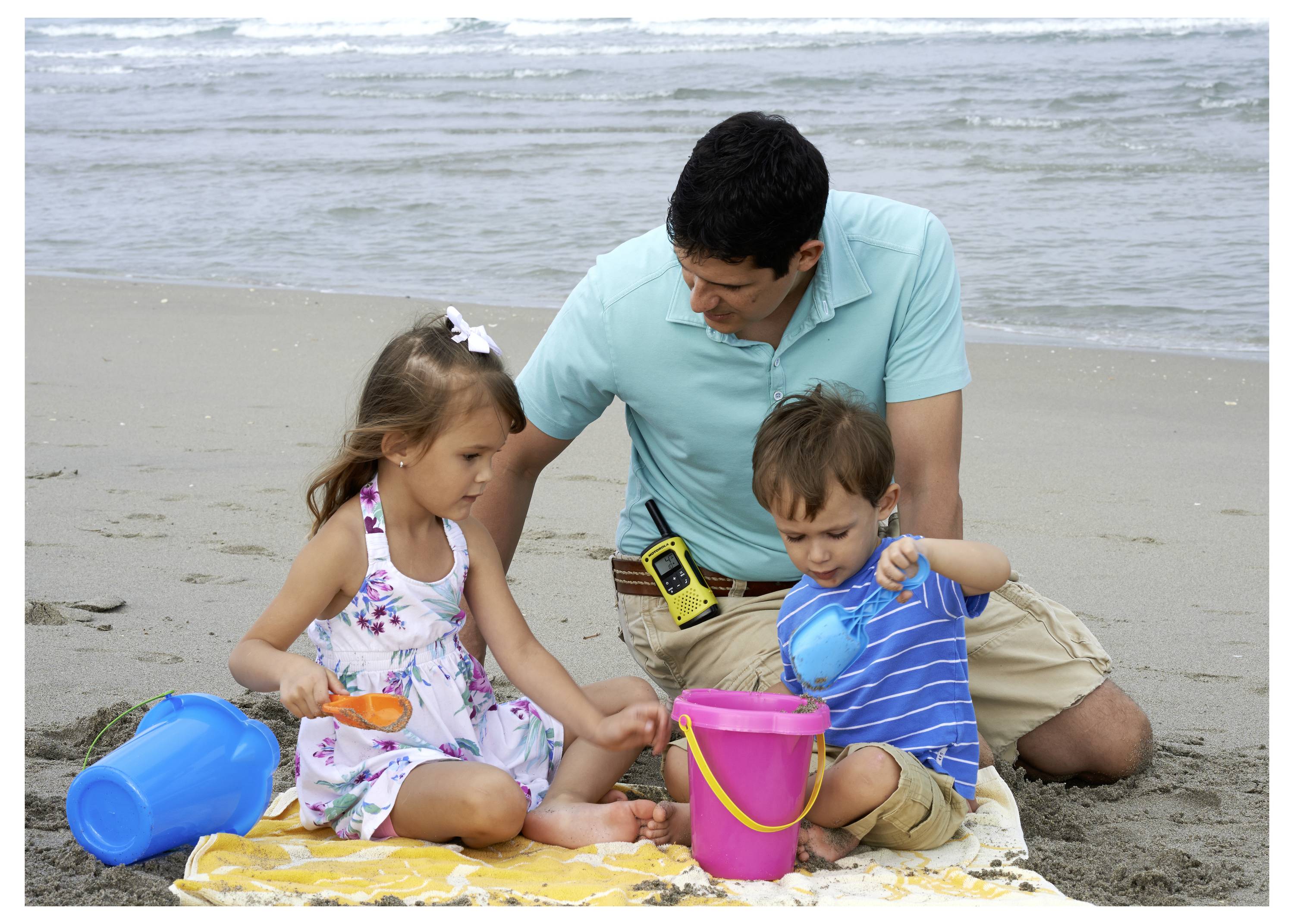 Un homme s'agenouille sur une plage avec deux enfants qui jouent dans le sable en utilisant des seaux et des pelles colorés près de l'océan.