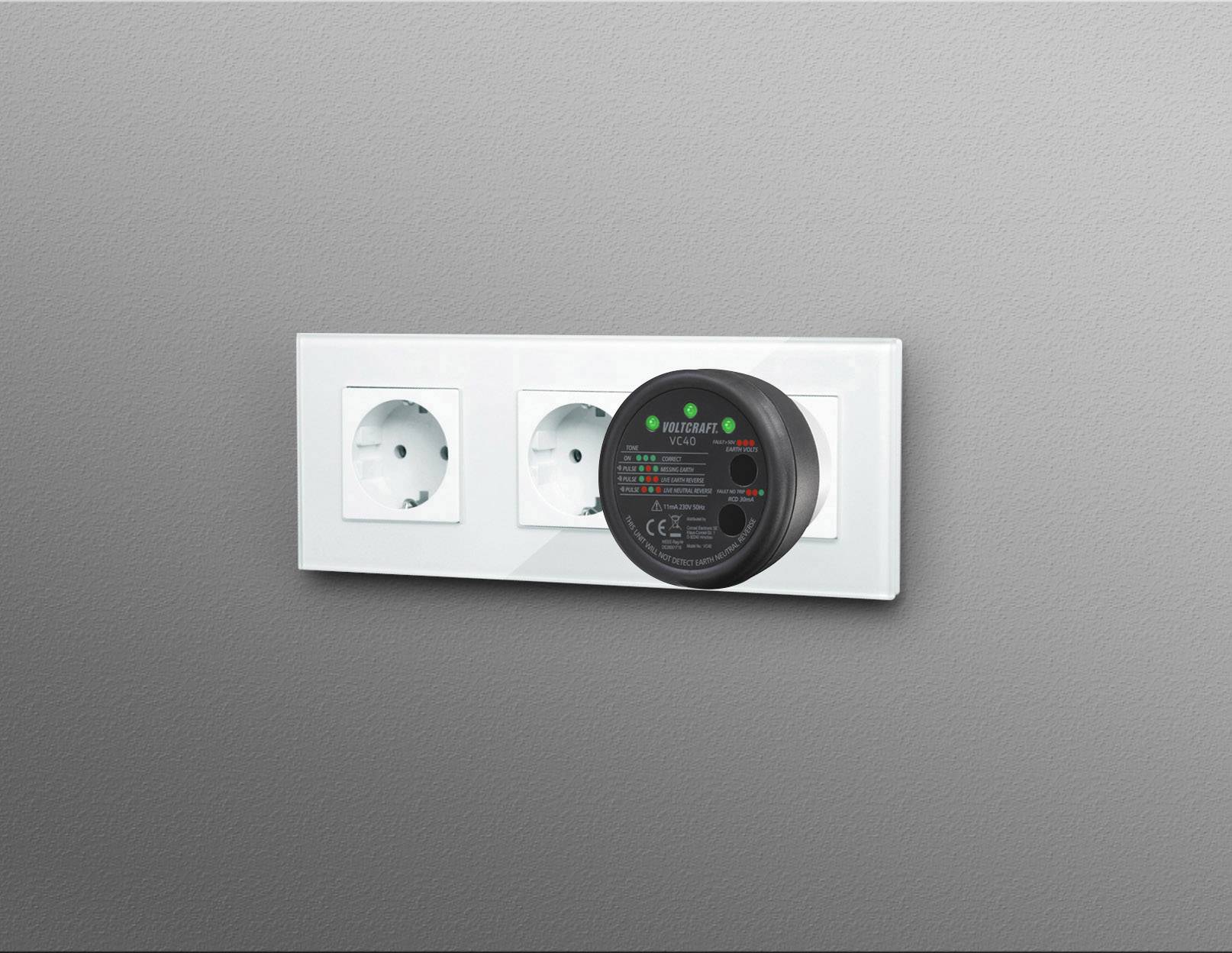 Trois prises électriques blanches sur un mur gris, avec un appareil de mesure numérique branché dans la prise de droite.