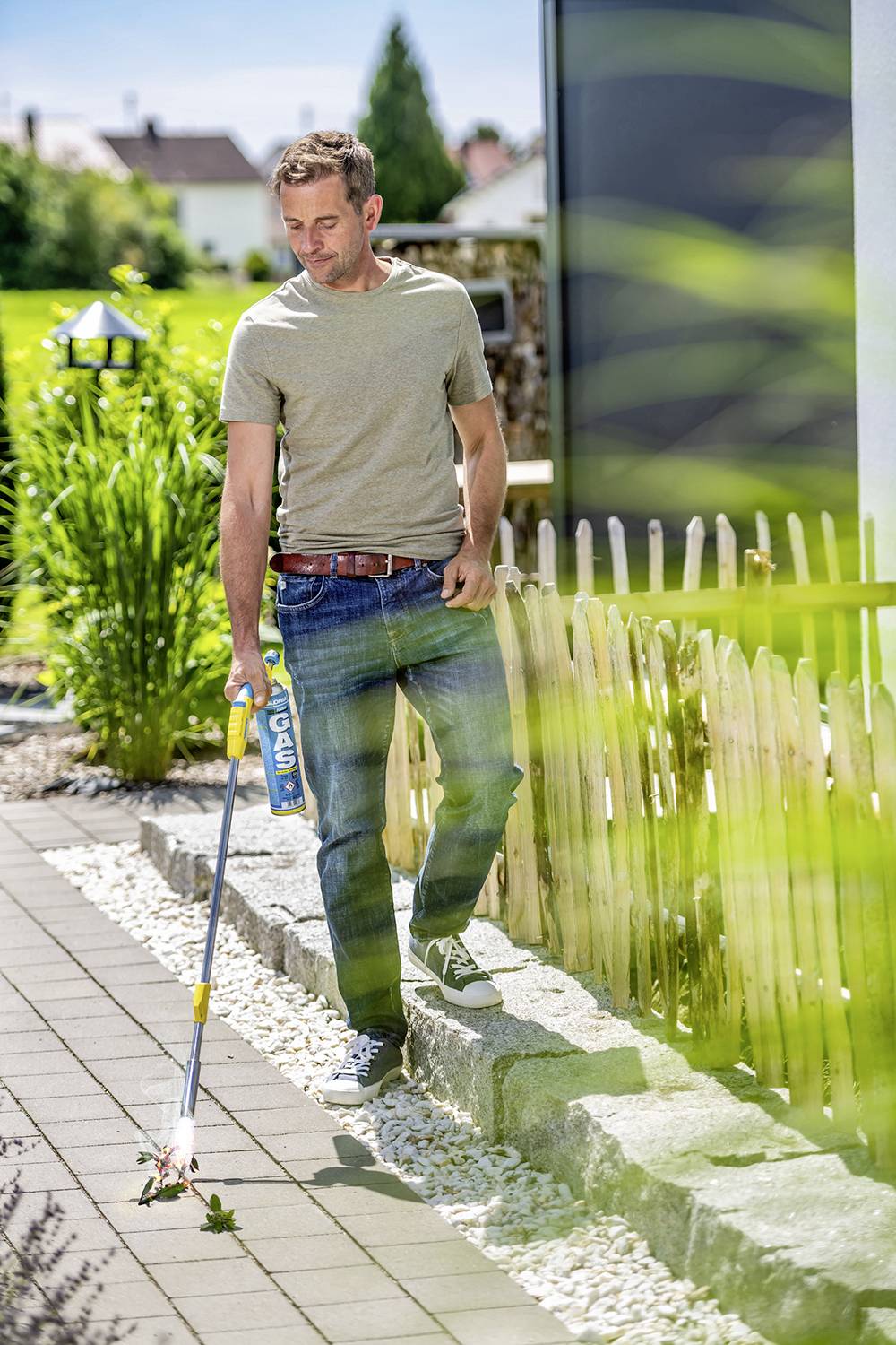 Un homme marche sur un chemin pavé dans un jardin et utilise un désherbeur thermique pour éliminer les mauvaises herbes.