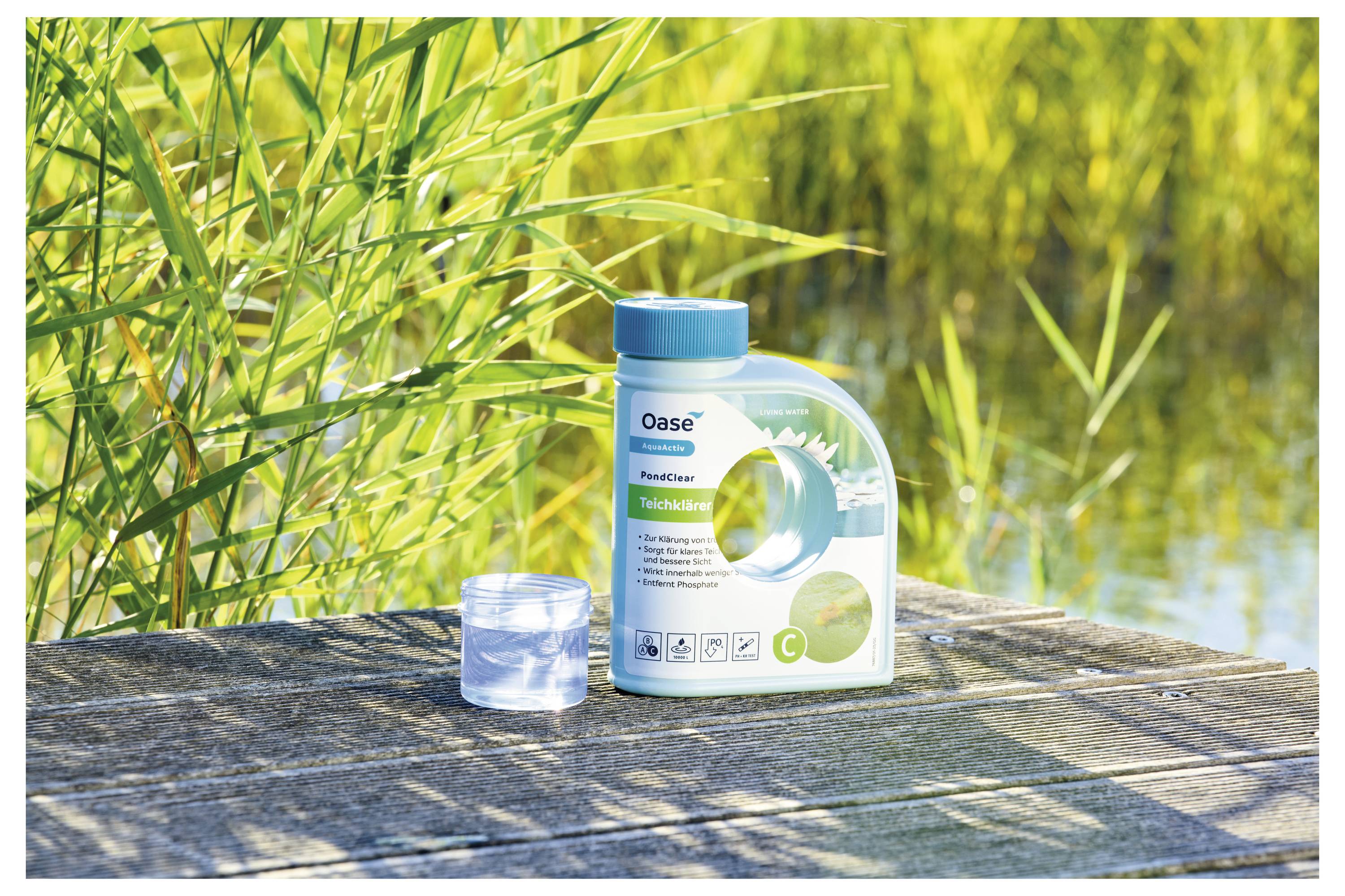 Un récipient de 'Oase PondClear' sur une surface en bois à côté d'une tasse à mesurer transparente. L'arrière-plan présente des roseaux verts luxuriants et de l'eau.