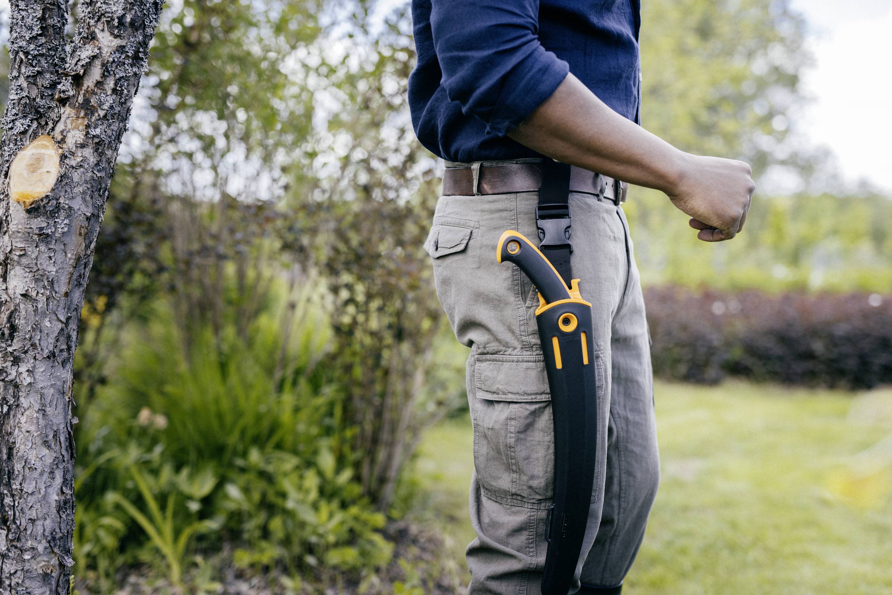 Un homme se tient dans le jardin, un couteau à sa ceinture, prêt pour les travaux de jardinage. En arrière-plan, on aperçoit des arbres et une pelouse.