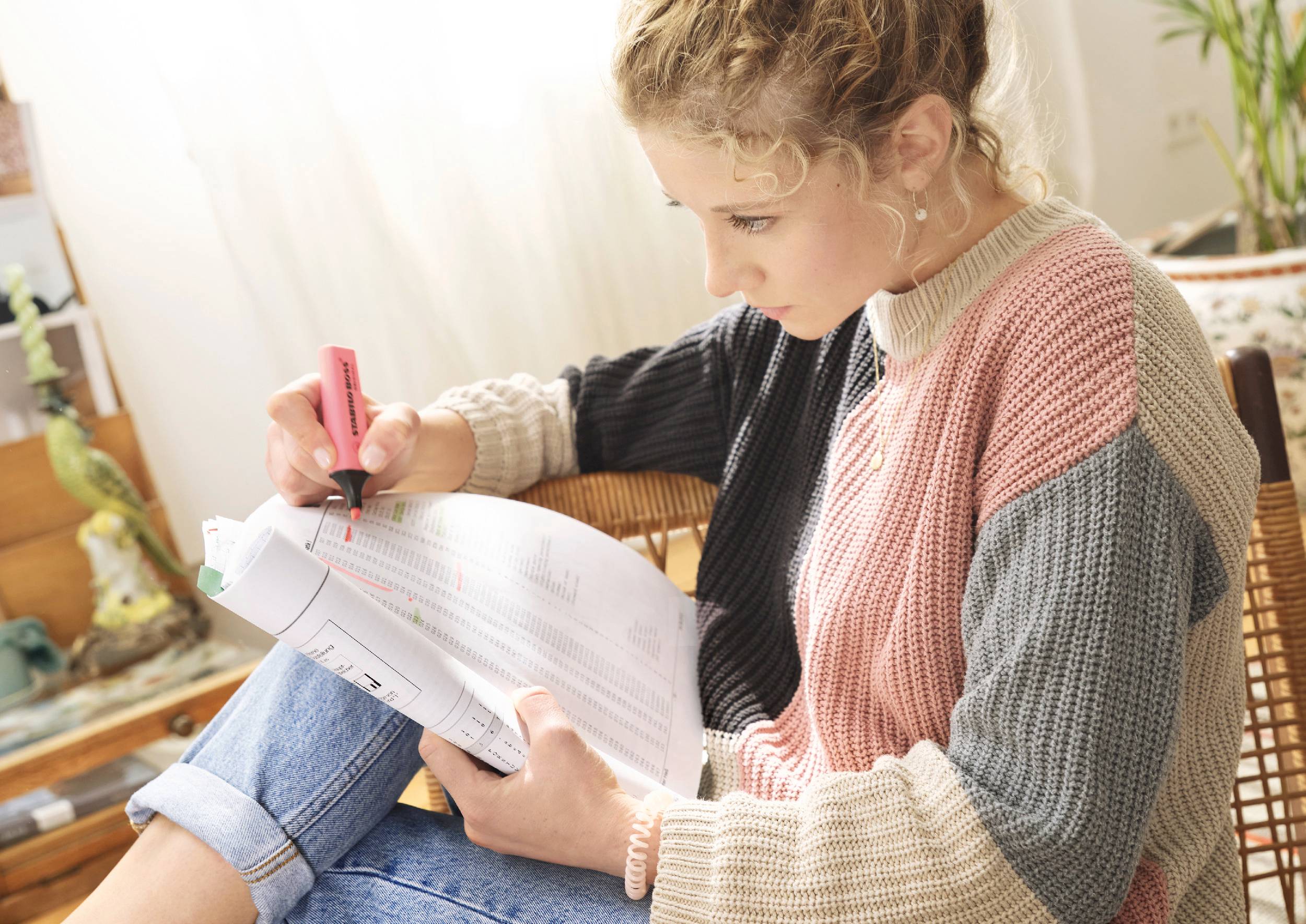 Une femme est assise dans un fauteuil et surligne des passages dans un document à l'aide d'un surligneur. Elle est vêtue d'un haut en tricot coloré.