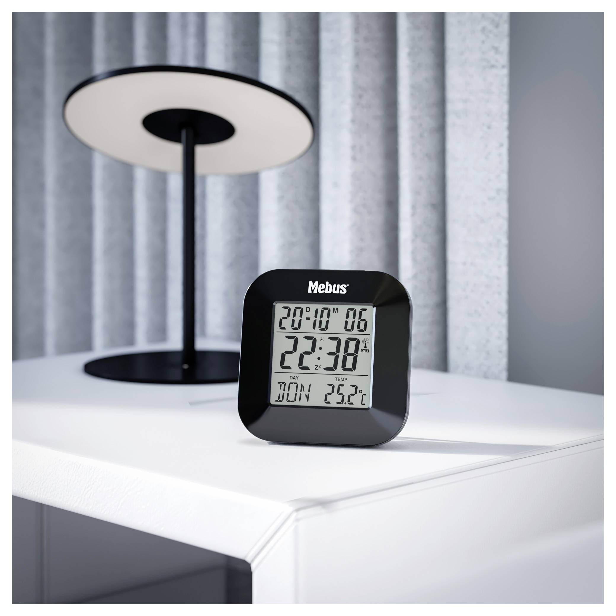 Une horloge numérique affichant « 20:10 », « 22:38 » avec « Dim 25°C » à côté d'une lampe sur une table blanche.