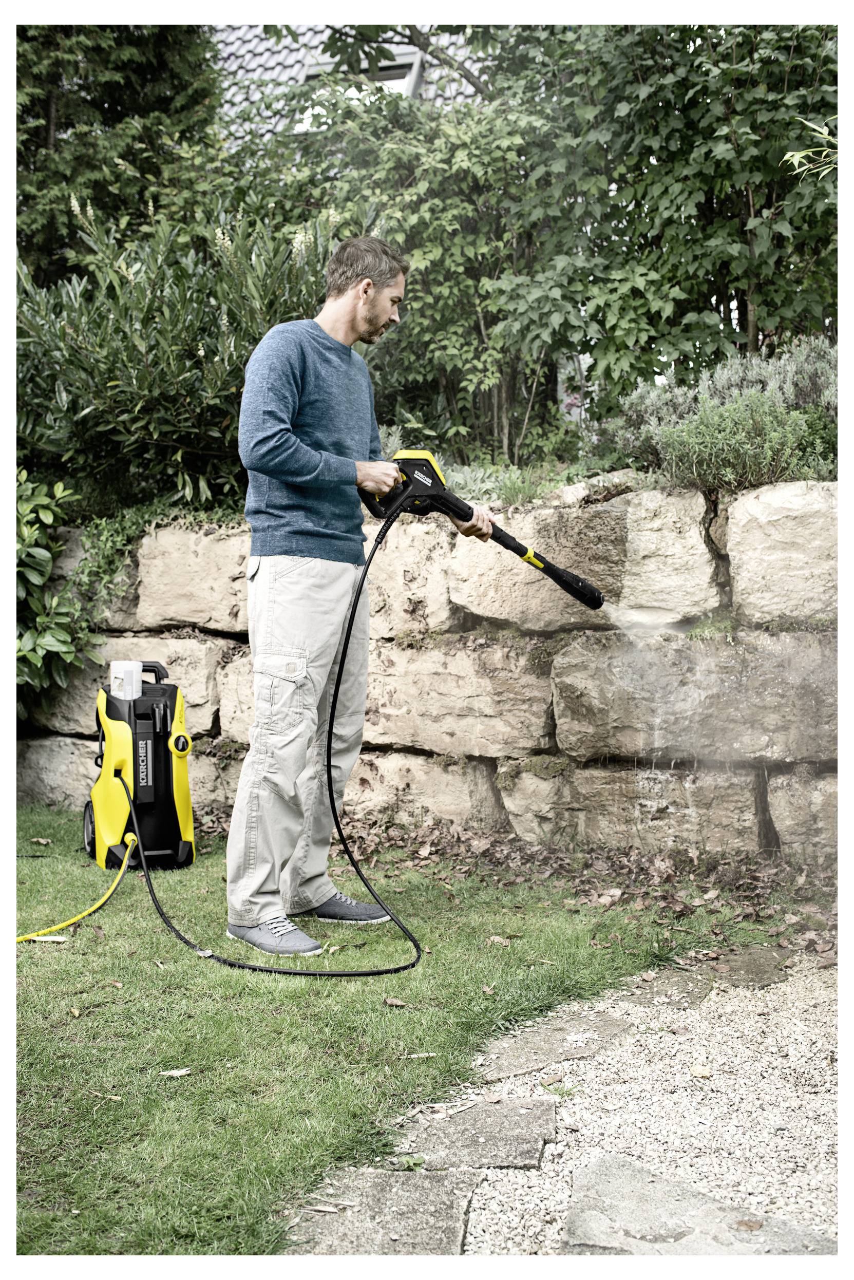 Une personne utilise un nettoyeur haute pression pour nettoyer un mur en pierre dans un jardin, entouré de verdure et avec une maison visible en arrière-plan.