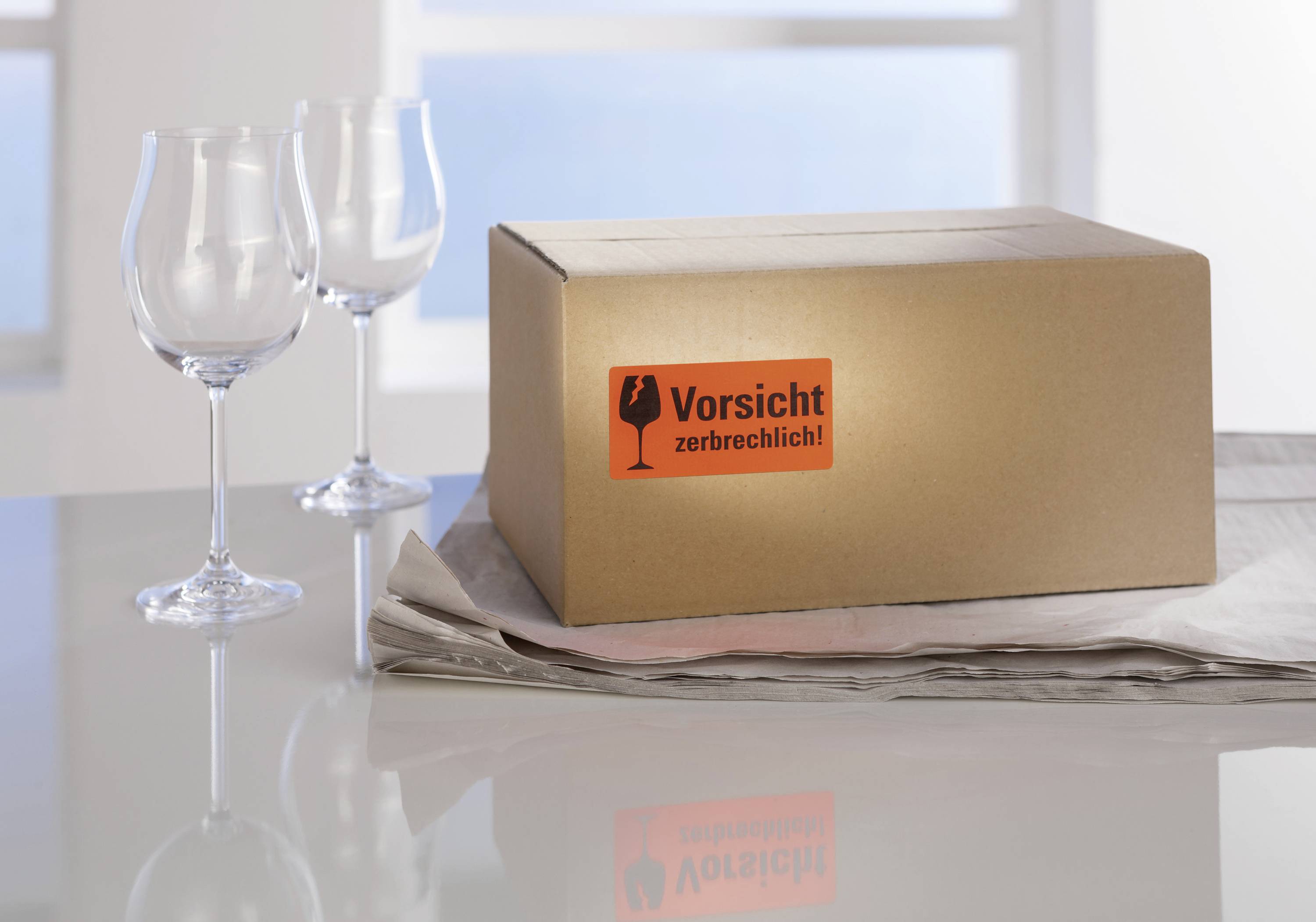 Un colis portant l'étiquette « Fragile » est posé sur une table avec deux verres à vin vides et du papier journal.