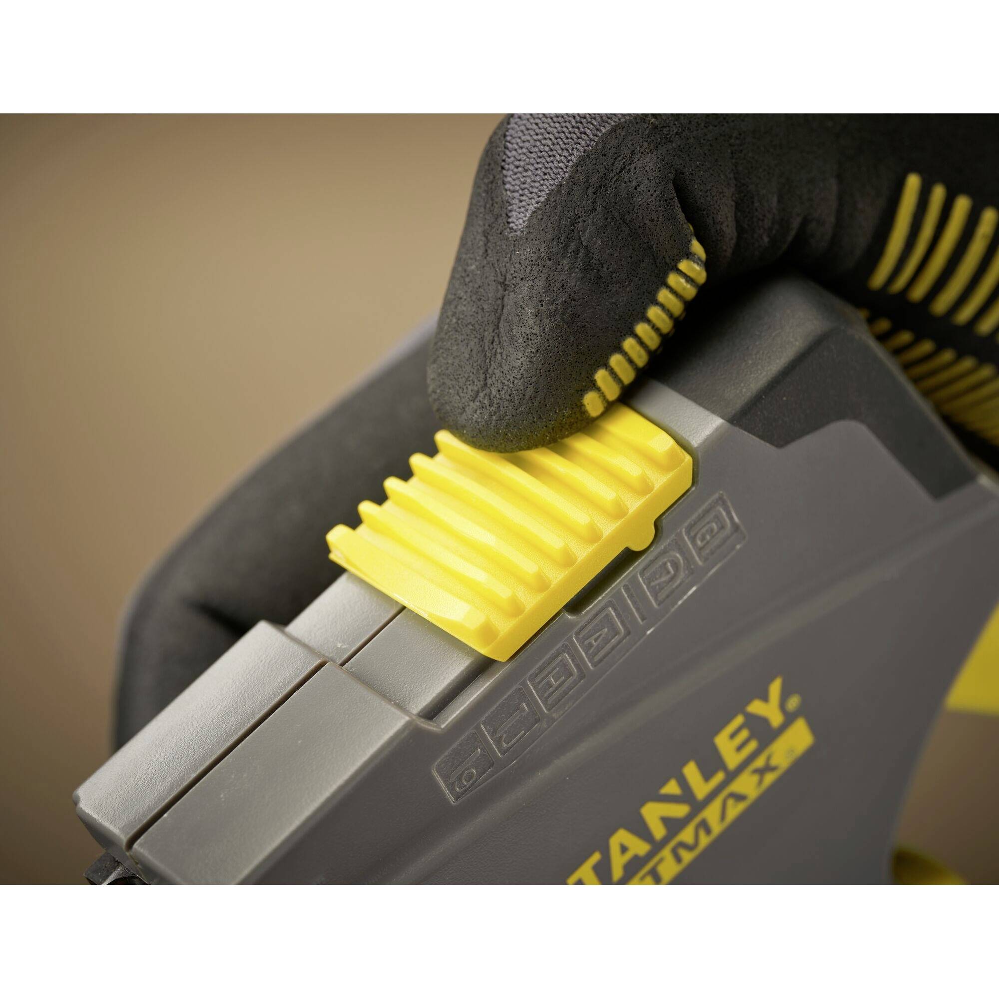 Une main portant un gant jaune tient un agrafeuse grise de la marque Stanley FatMax et appuie sur le bouton de déclenchement jaune.