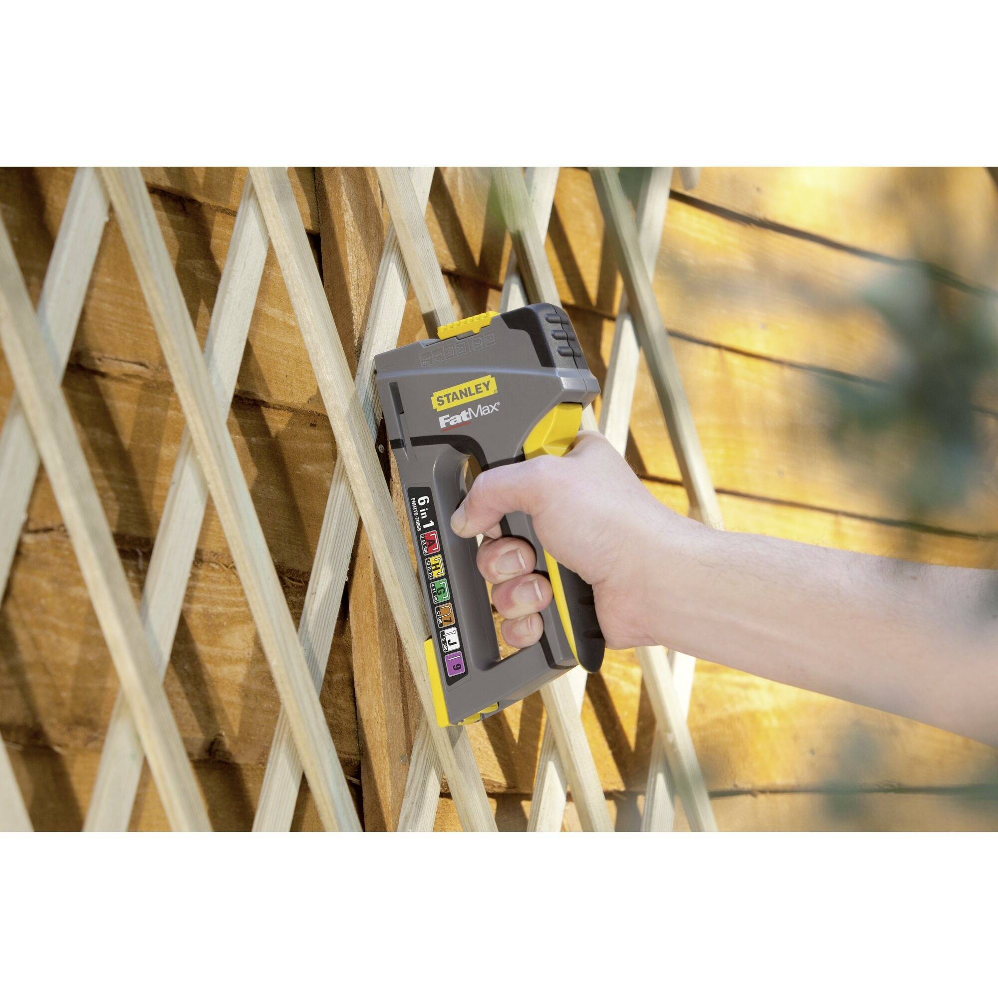 Une main utilise un pistolet à agrafes jaune pour fixer un filet grillagé à une clôture en bois.