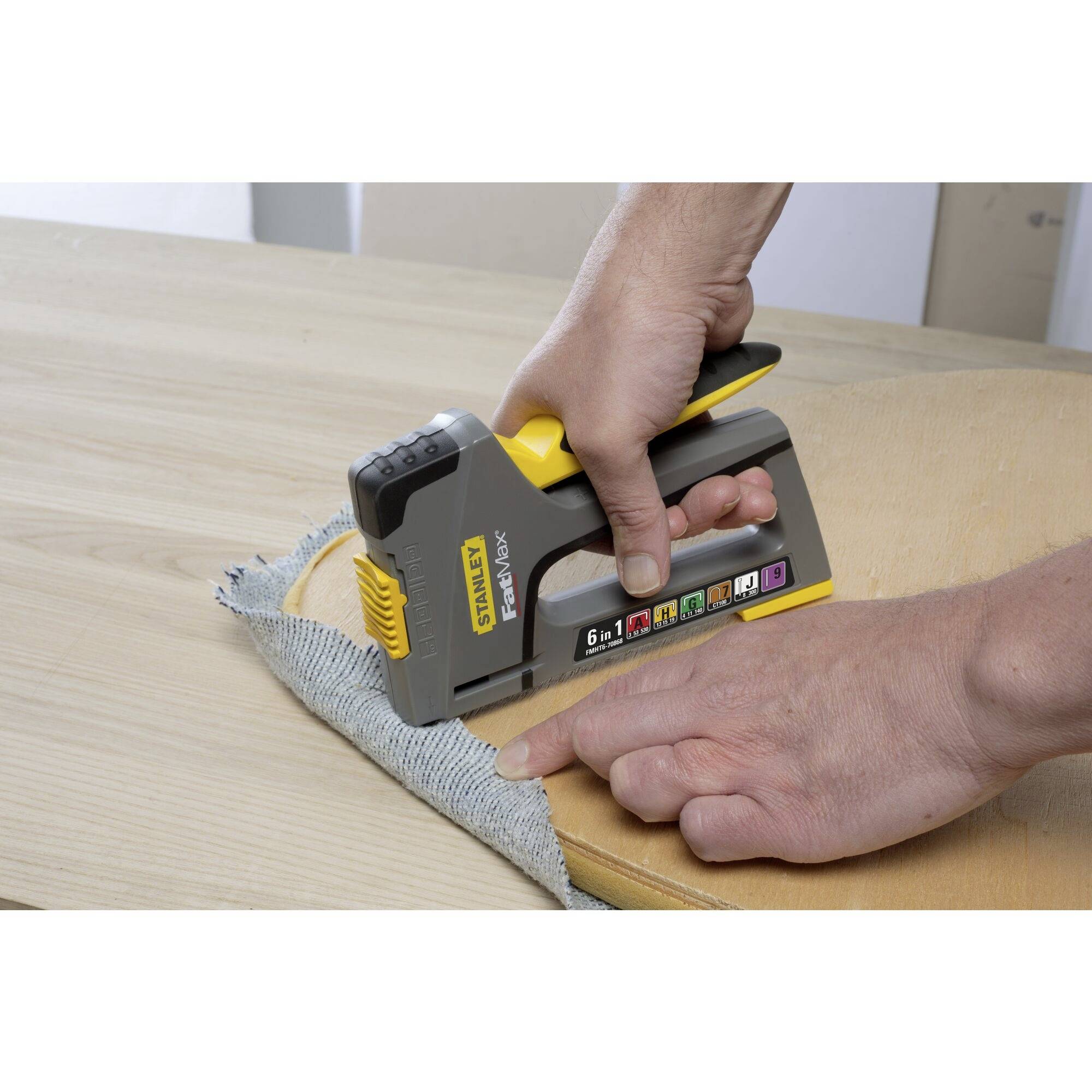Une personne tient un pistolet à agrafes et fixe un morceau de tissu sur une surface en bois. Le pistolet à agrafes est gris et jaune.