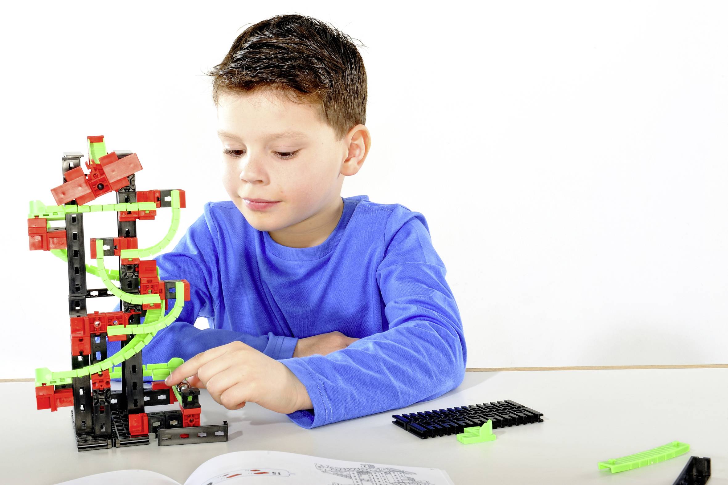 Un garçon portant un pull bleu joue avec concentration à un kit de construction, composé de blocs rouges et verts, posé sur une table.