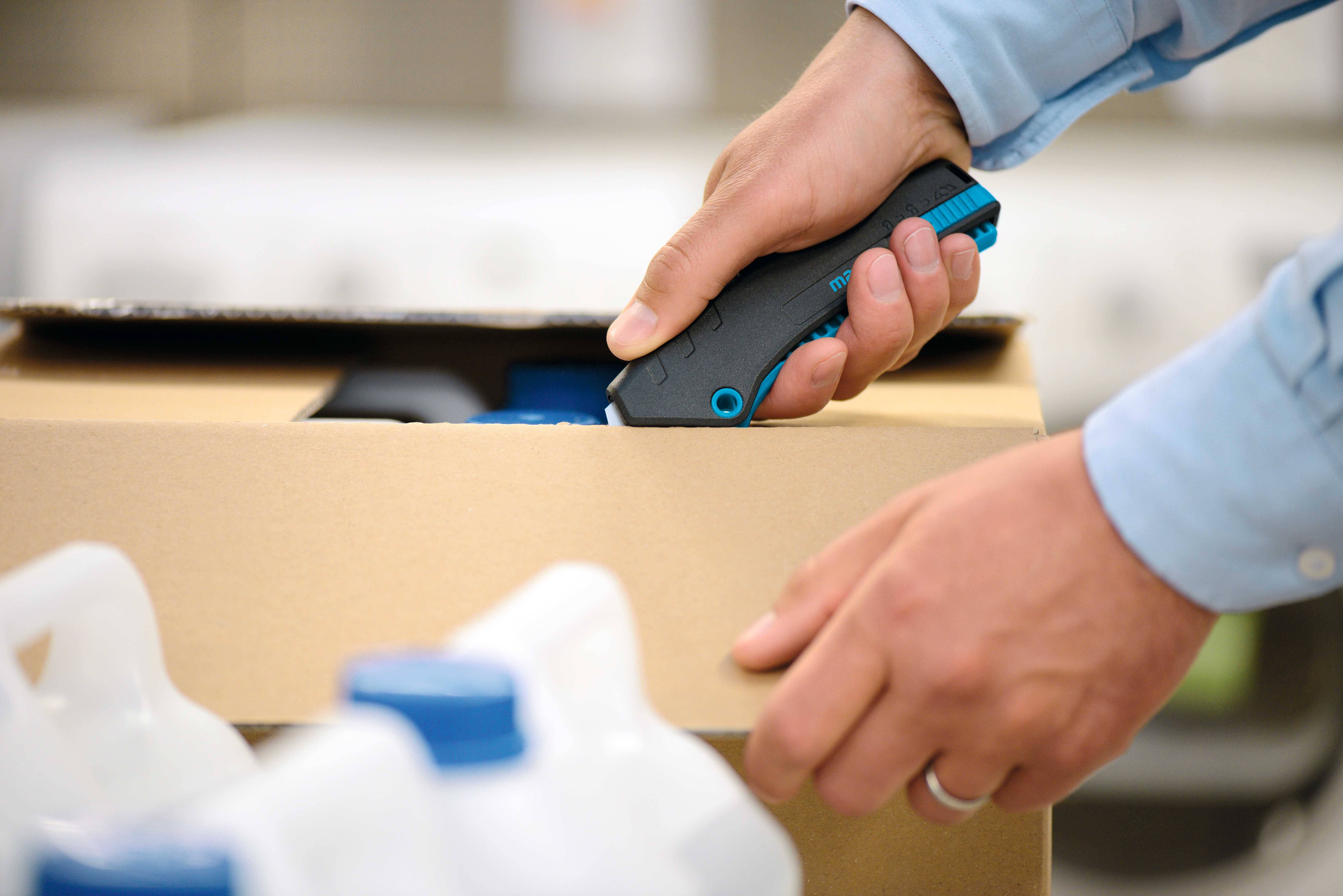 Une personne ouvre un carton en carton à l'aide d'un cutter. À côté du carton se trouvent des contenants blancs en plastique avec des bouchons bleus.