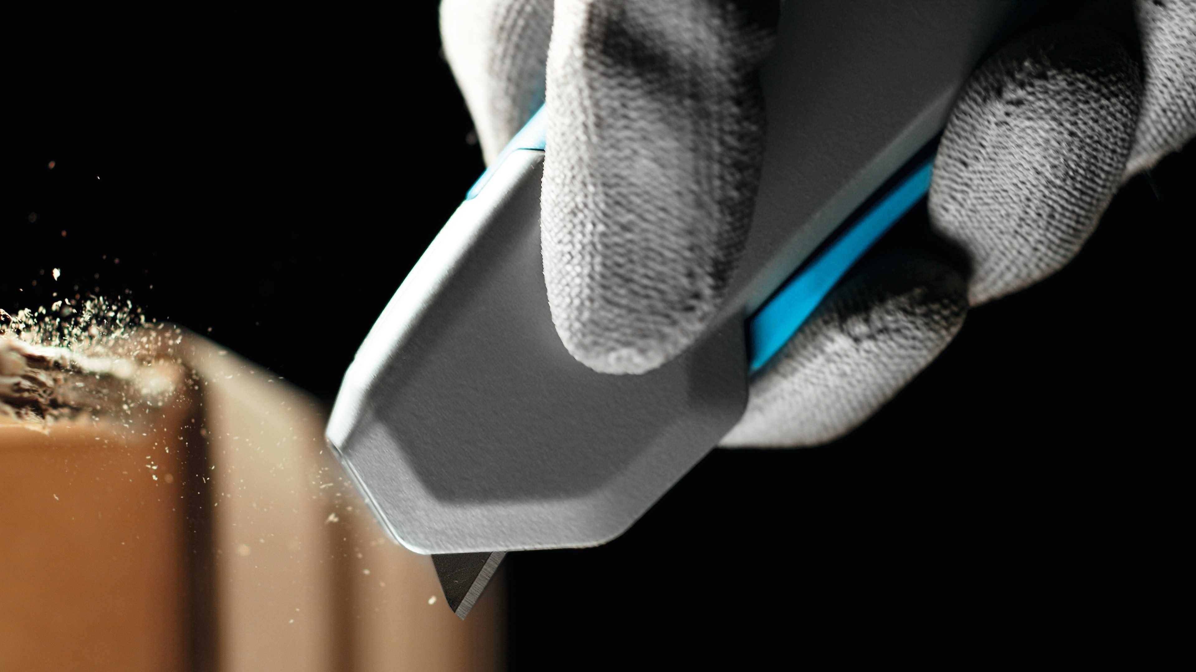 Une main gantée de gris tient un cutter et coupe précisément un morceau de carton, des copeaux de bois étant visibles.