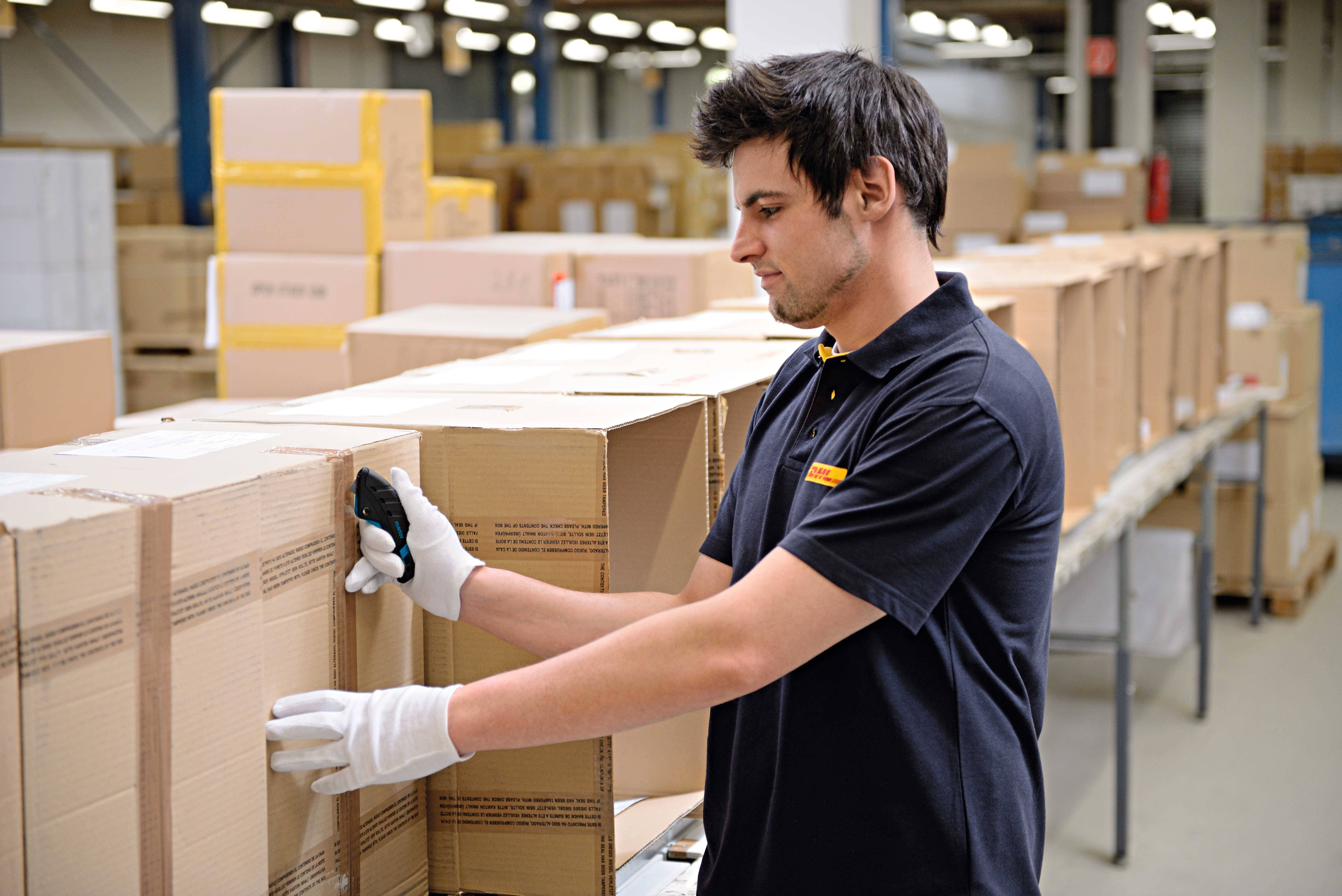 Un homme scanne des colis dans un entrepôt. Il porte des gants et des vêtements de travail. À l'arrière-plan, de nombreuses boîtes sont visibles.