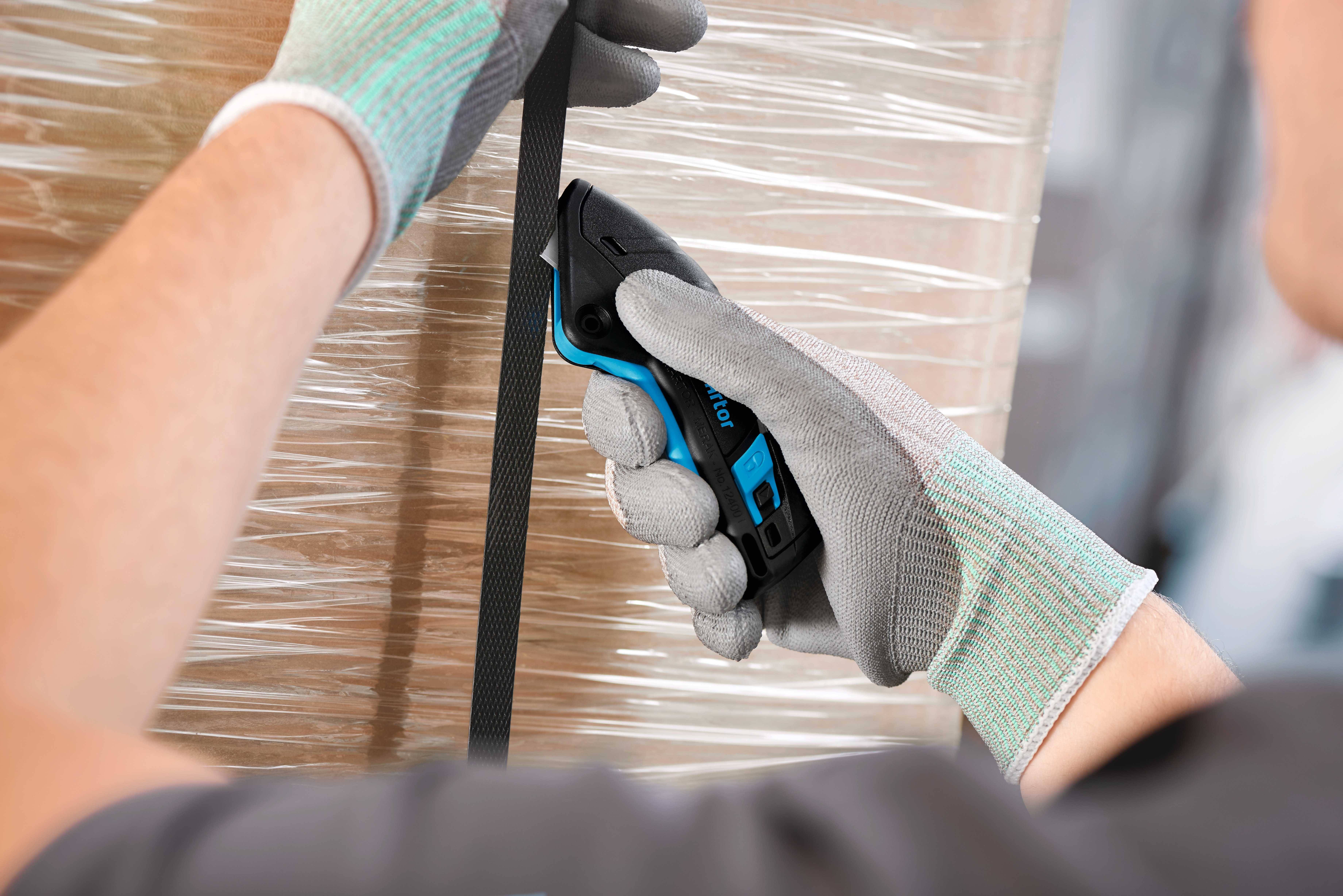 Une personne portant des gants gris utilise un couteau de sécurité pour couper un bandeau noir en plastique autour d'un emballage.