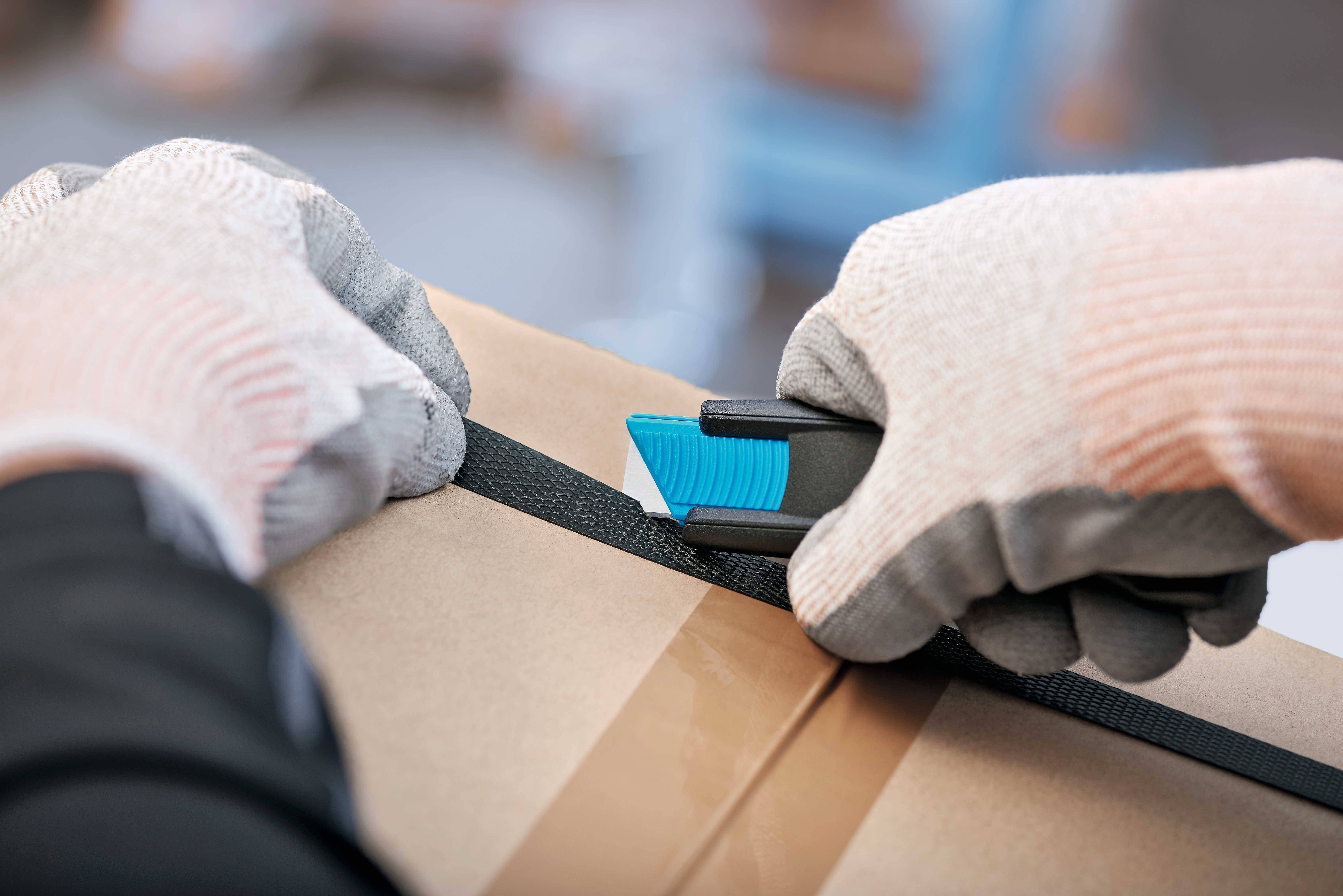 Une personne portant des gants coupe un ruban noir autour d'un carton à l'aide d'un cutter bleu.