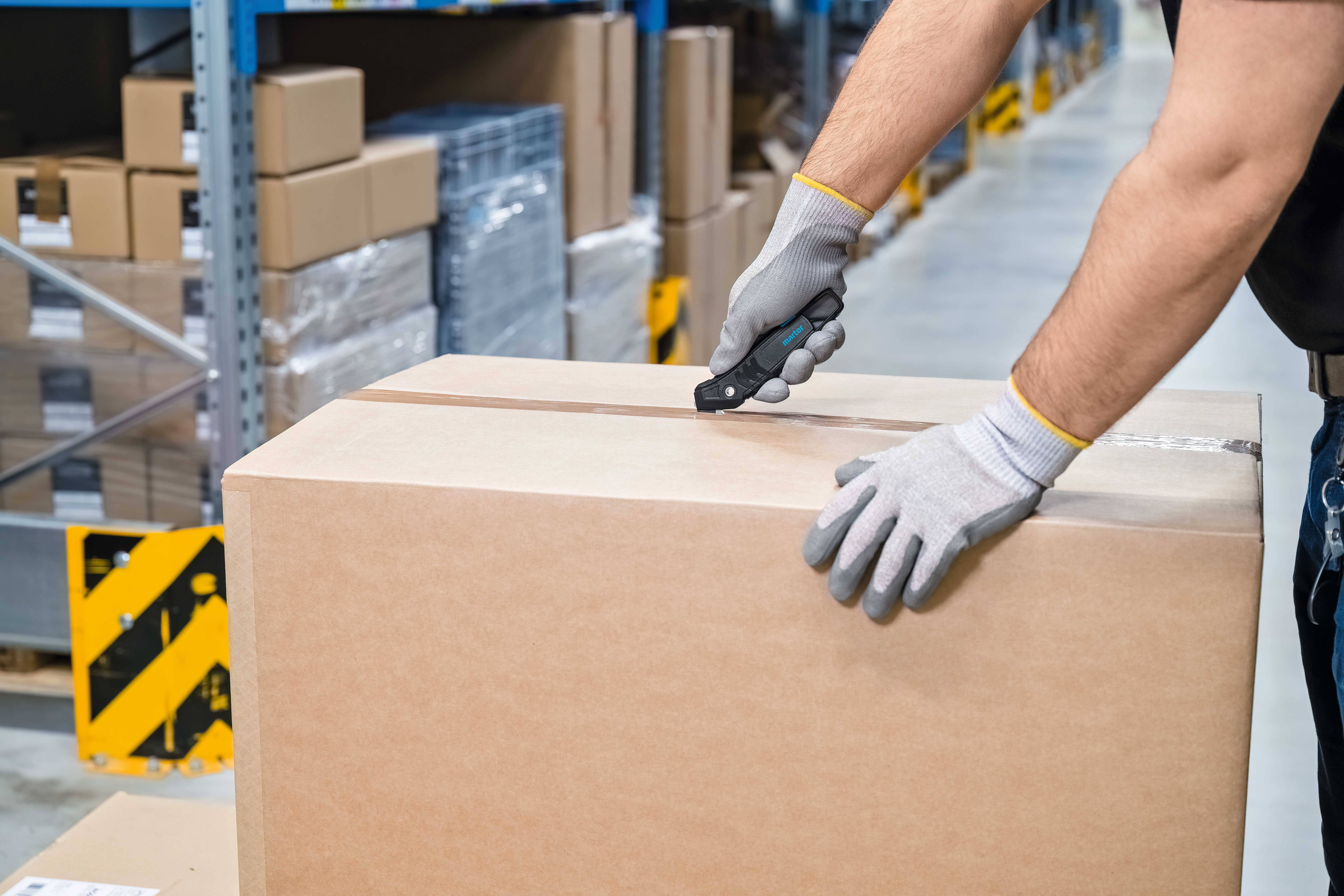 Une personne portant des gants de travail ouvre un grand carton à l'aide d'un couteau dans un entrepôt avec des étagères remplies de colis.